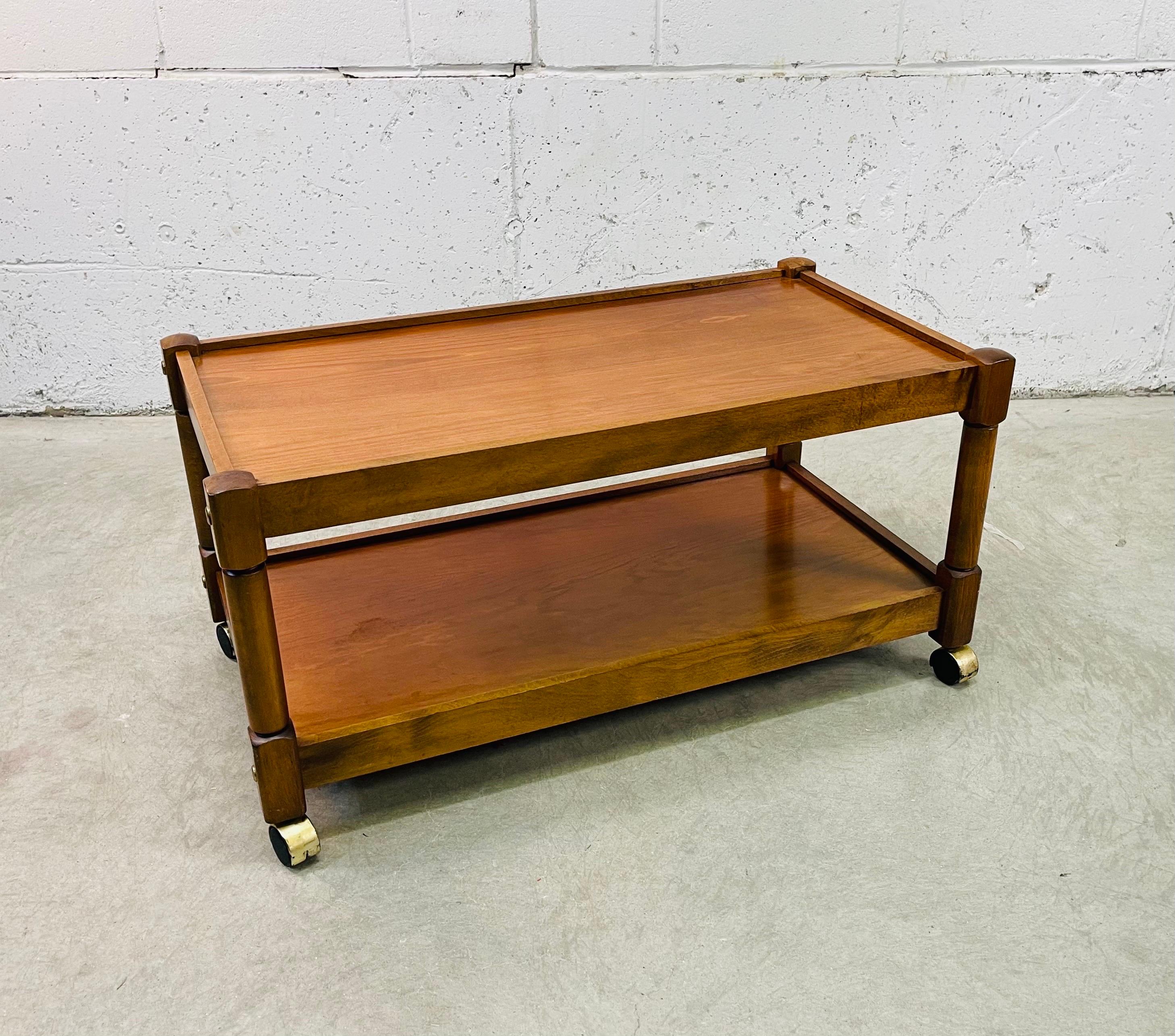 Vintage 1960s two-tier mahogany rectangular rolling serving cart. The cart has two shelves for storage and rolls freely. The cart has gold metal accents. Marked Romania.