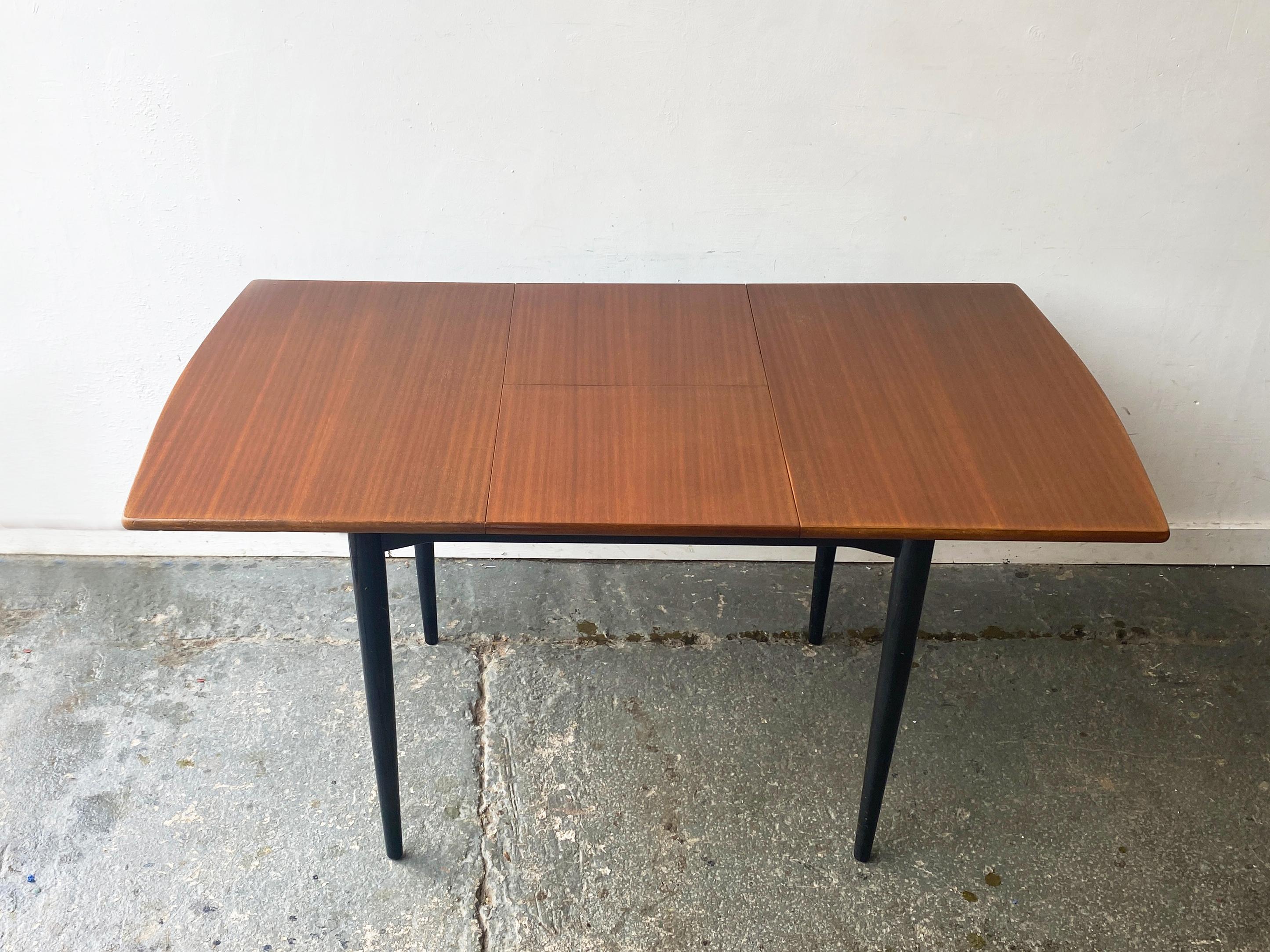 A compact mid-century modern extending dining table by White & Newton, who were renowned for their high-quality craftsmanship. The table has a teak top with gently curved edges, and is supported by tapering black legs. It extends by using central