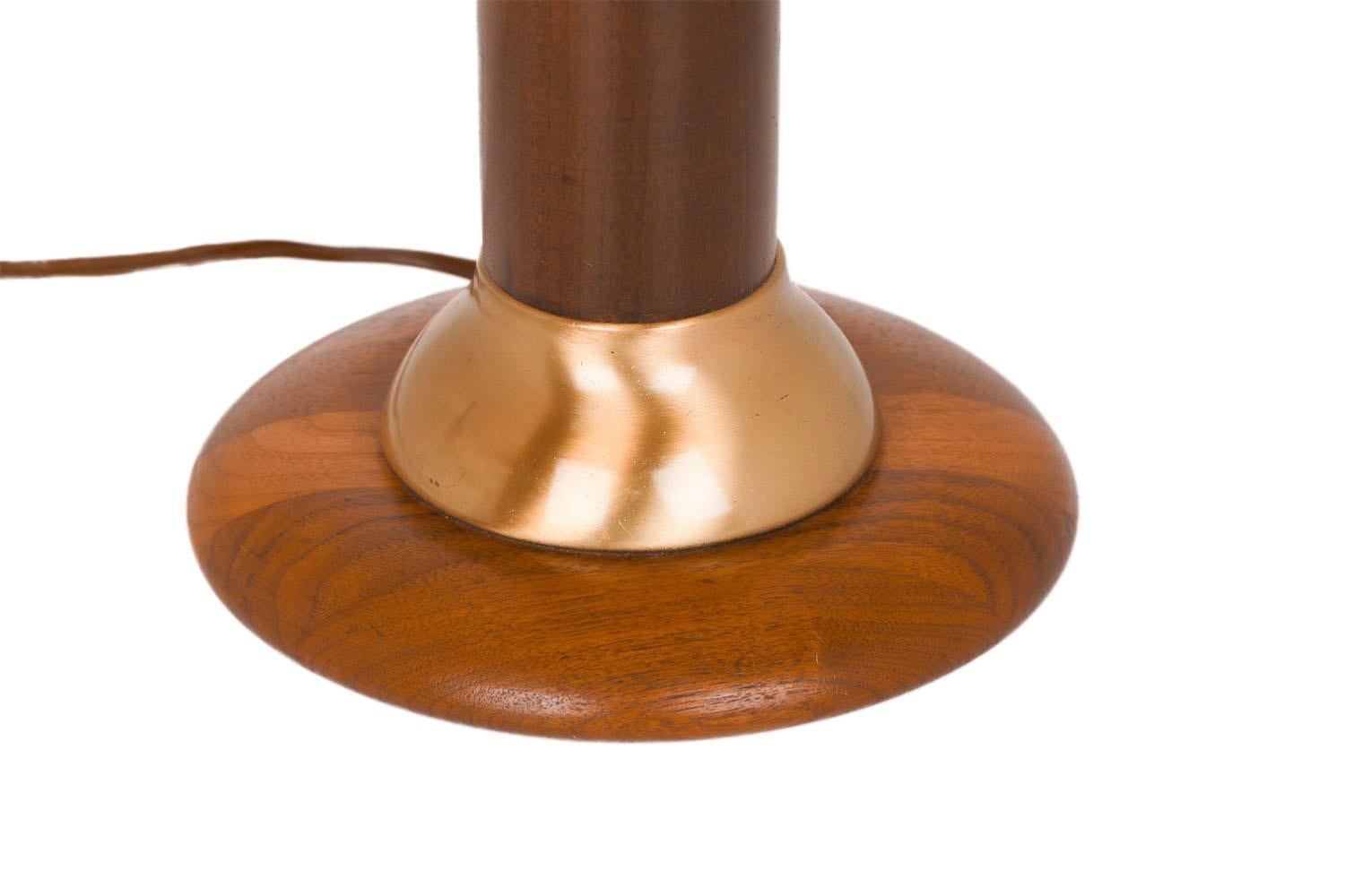 Lampada da tavolo Mid-Century Modern del 1960 in noce tornito e ottone con paralume originale. La base in legno tornito, alta, sottile e modernista, è separata dal supporto a disco in noce con un'ampia fascia in ottone curvato. Il paralume originale