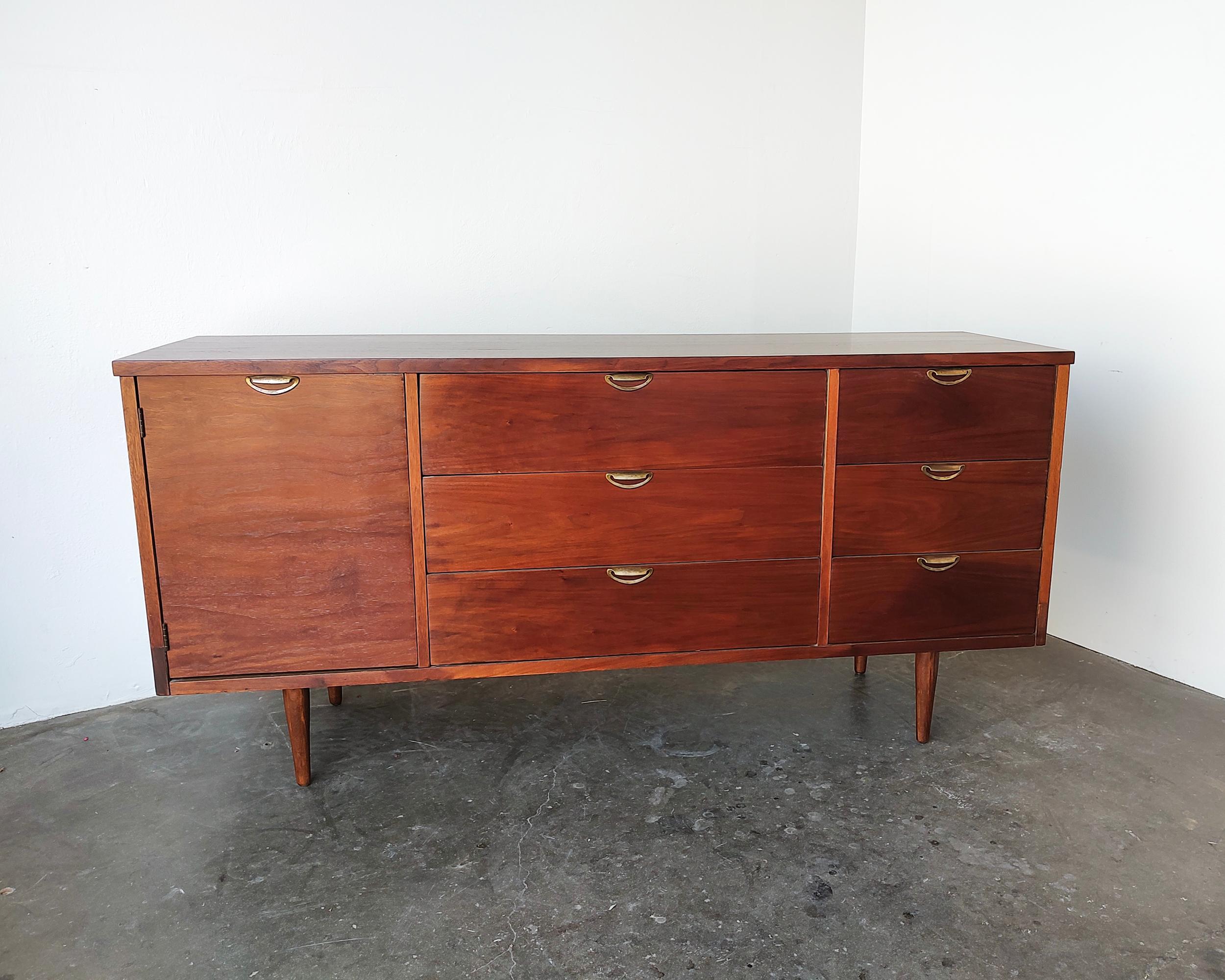 Beautiful restored mid-century modern walnut wood sideboard cabinet by Bassett Furniture Inc. circa 1960s. Six drawers on the right (one with built-in dividers) and cabinet on the left with removeable shelf featuring original patinaed brass pulls.