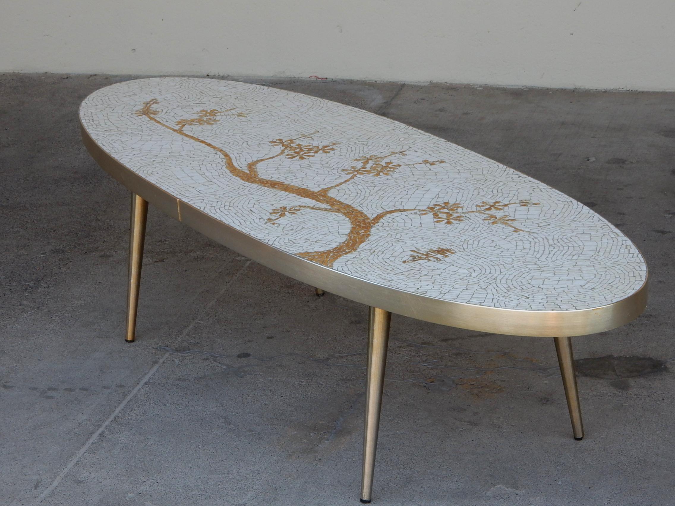 Fabulous mosaic art coffee table attributed to Berthold Muller.
Surfboard shape banded in gold with matching legs.
Bone white tile mosaic with gold leaf bonsai design.
Not signed, circa 1950s.