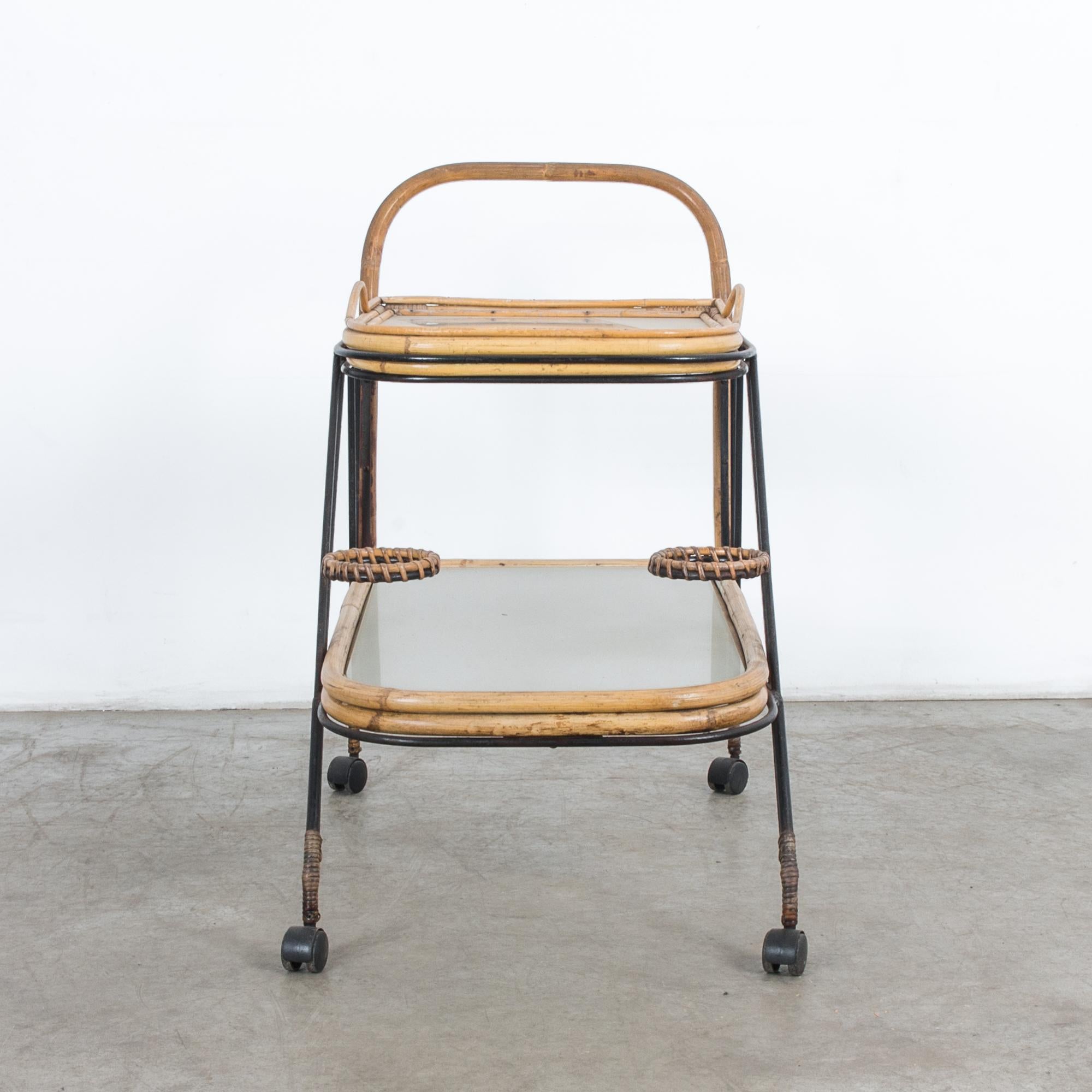 A wheeled cart from France, circa 1960. A slender metal frame on wheels supports two tiered tabletops made of bamboo and opaque glass. A bamboo handle permits the cart to be pushed, while handles on the upper table allow it to be lifted as a tray.