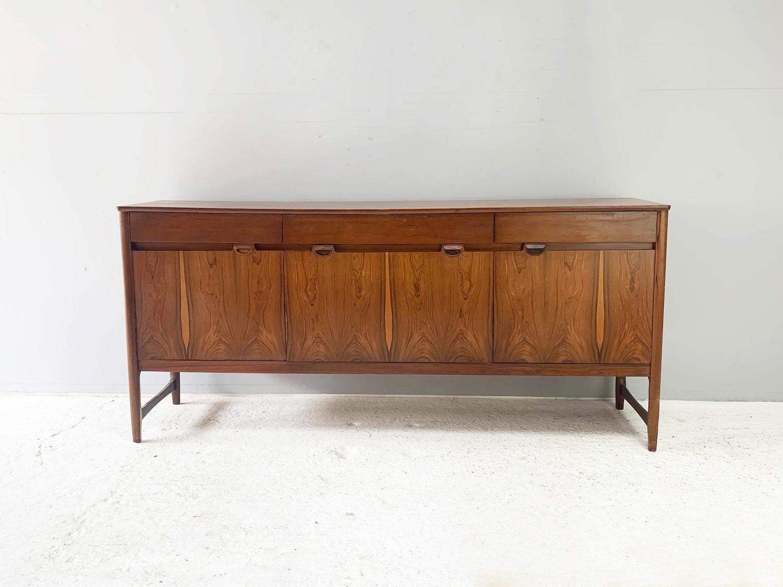 A rare Rosewood and solid teak sideboard by respected British maker Nathan Furniture, part of their premium ‘Caspian’ range.

Rectangular Rosewood top above three frieze drawers which are over a central fall flap drinks cabinet. Flanked by two