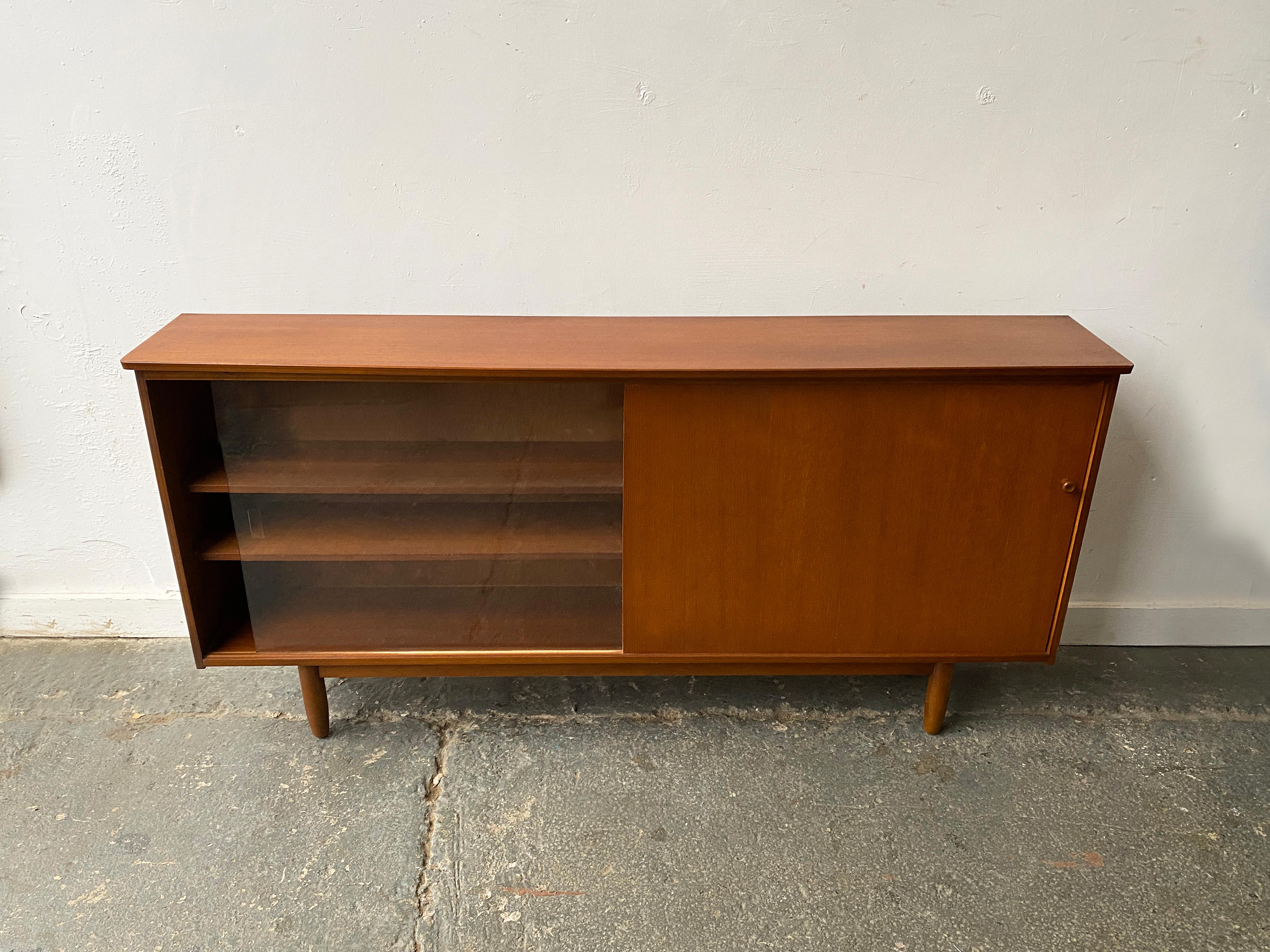 Ein Mid-Century Modern Bücherregal des englischen Herstellers Avalon mit 2 Schiebetüren, eine aus Holz, die andere aus Glas. Die Merkmale sind wie folgt:

- Rahmen aus Teakholz
- 1 Glas- und 1 Teakholz-Schiebetür
- Verstellbare Regale
- Von