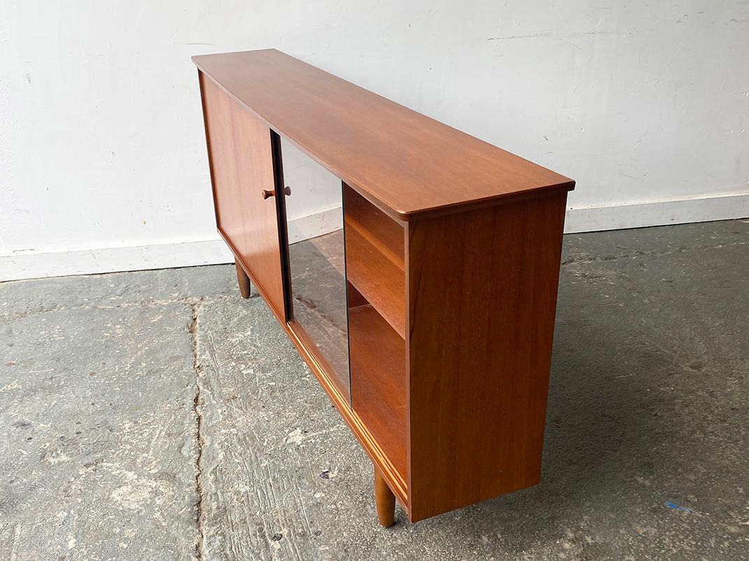 A Mid-Century Modern book case by English manufacturer Avalon with 2 sliding doors, one wood and the other glass. Features are as follows:

- Teak frame
- 1 glass and 1 teak sliding door
- Adjustable shelving
- By English maker: Avalon
- Long and