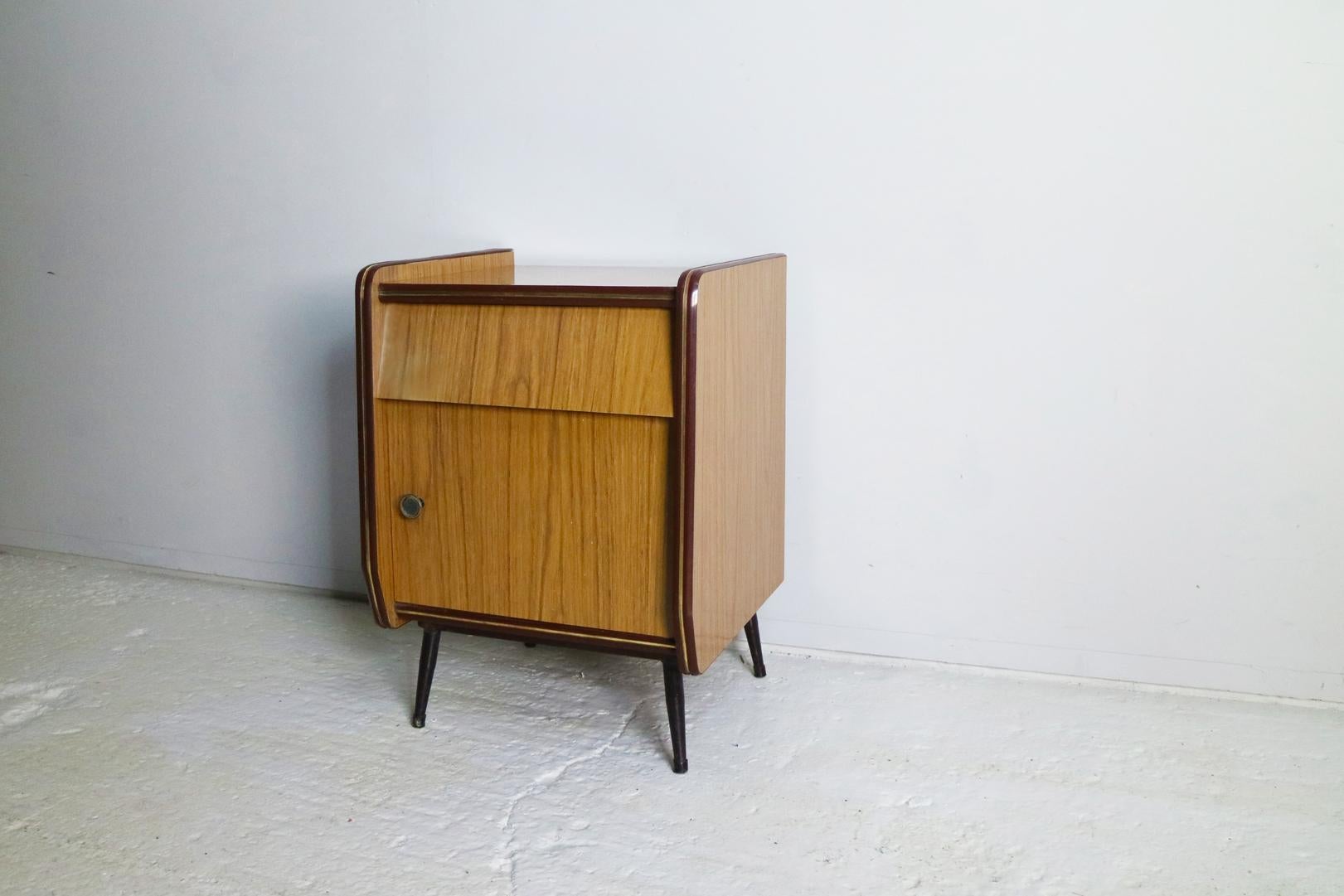 A 1960s single bedside cabinet with one drawer above a cupboard. Formica veneer throughout, finished with a maroon plastic and brass trim. Sits on painted metal legs.

  