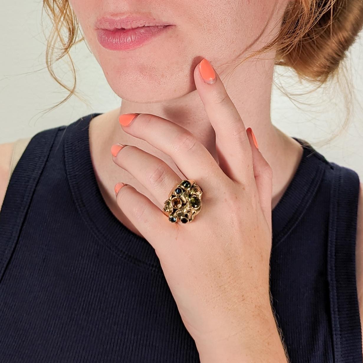 A fine Modernist ring.

By the New York City Modernist female goldsmith Resia Schor. 

In 14k gold with multi-gemstones.

Comprising a Brutalist head with varied gold elements and bezel-set gemstones. Set with an oval emerald cabochon, a green