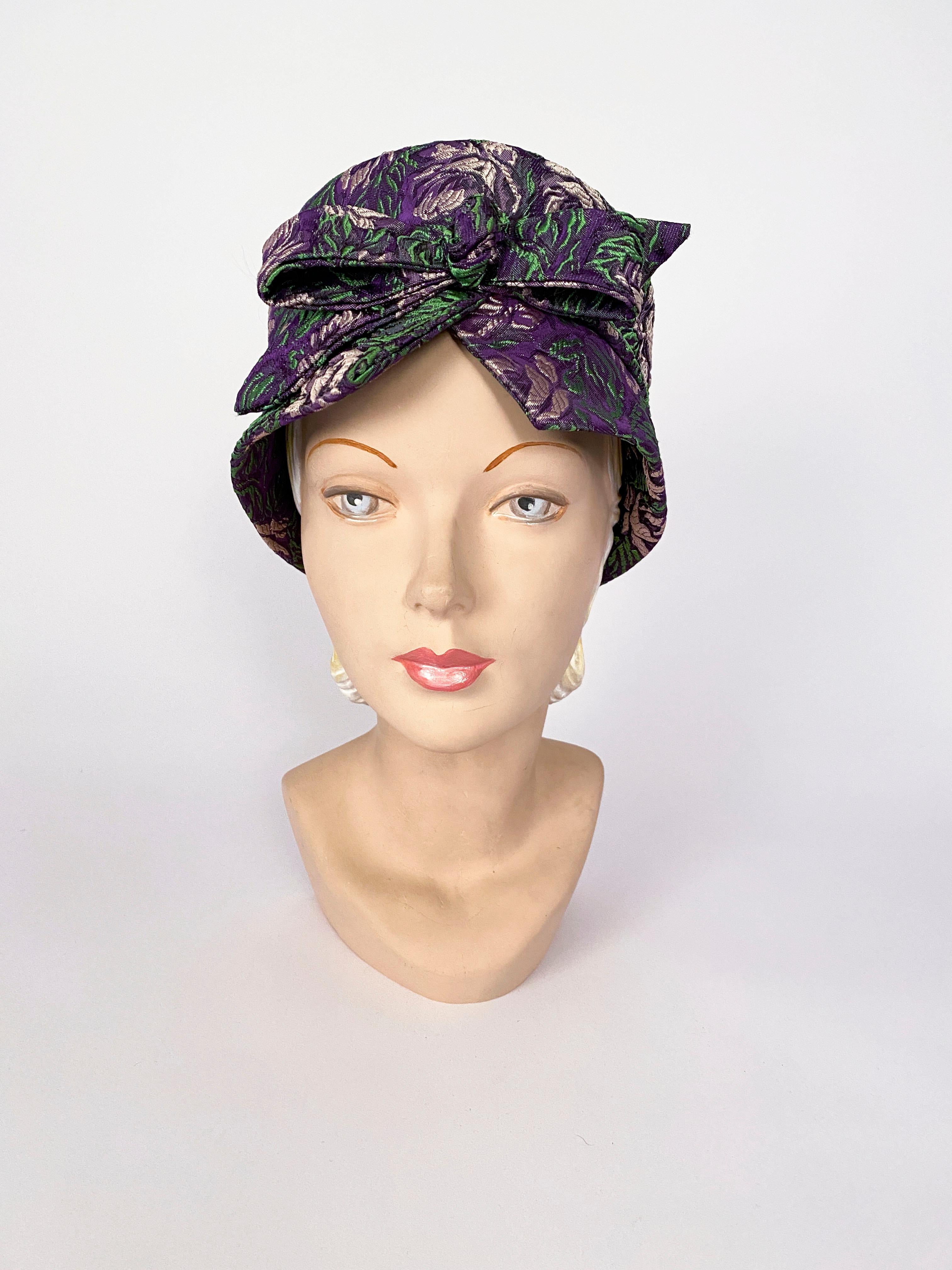 1960s Mr. John purple hat with a turban-like structure. The entire hat is made of a textured floral brocade with undertones of lavender and green. This hat is finished with a large tied bow.