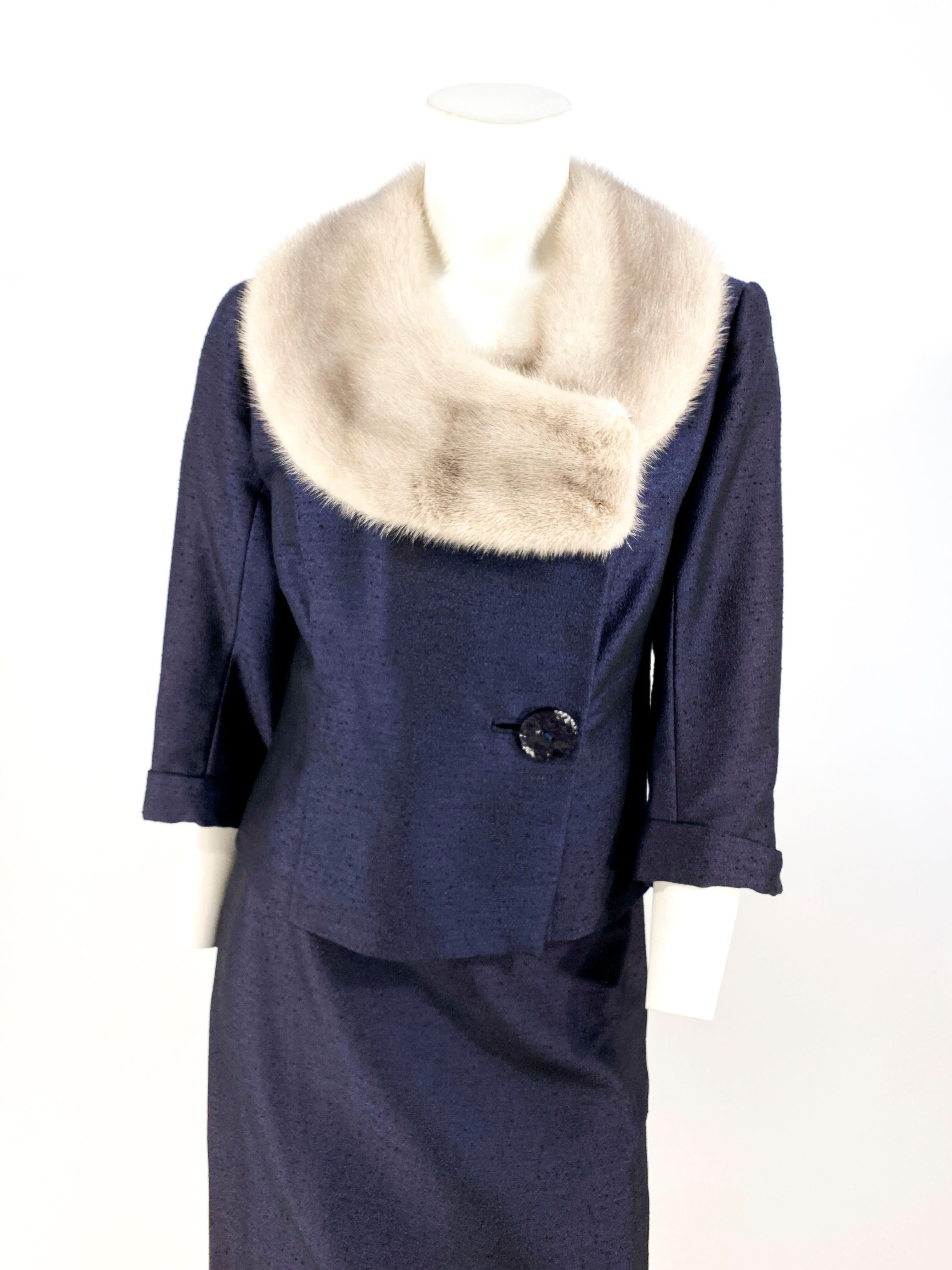 1960s navy blue textured suit with a cropped jacket featuring an enlarged silver grey mink shawl collar, large designer buttons, three-quarter length cuffed sleeves, and a asymmetrical jacket face. The jacket is fully lined but the straight skirt is