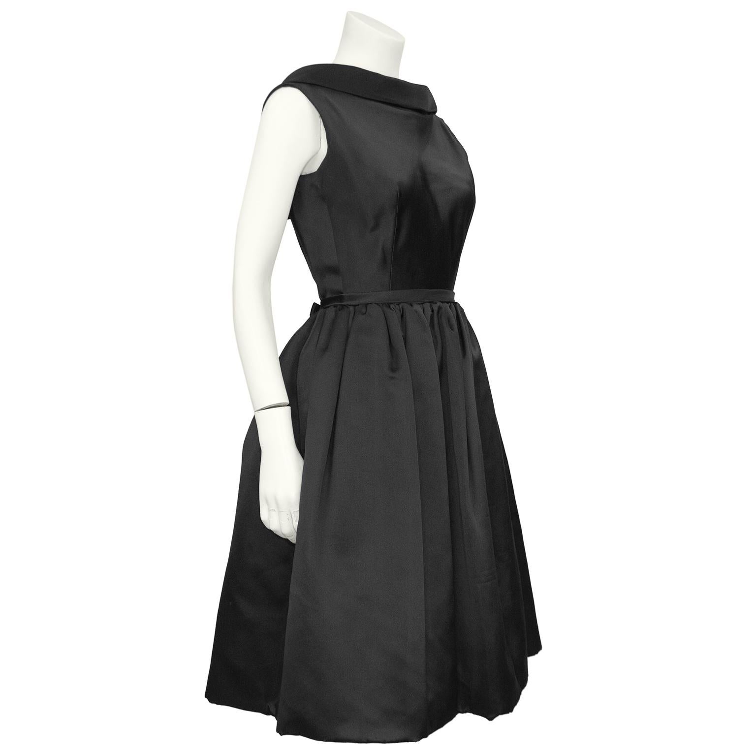 Classic and stunning black silk satin Norman Norell dress from the 1960s. Sleeveless with an oversized folded shawl collar. Fitted through the body with four round fabric covered buttons. Thin belt at waist that can be tied into a perfect bow that