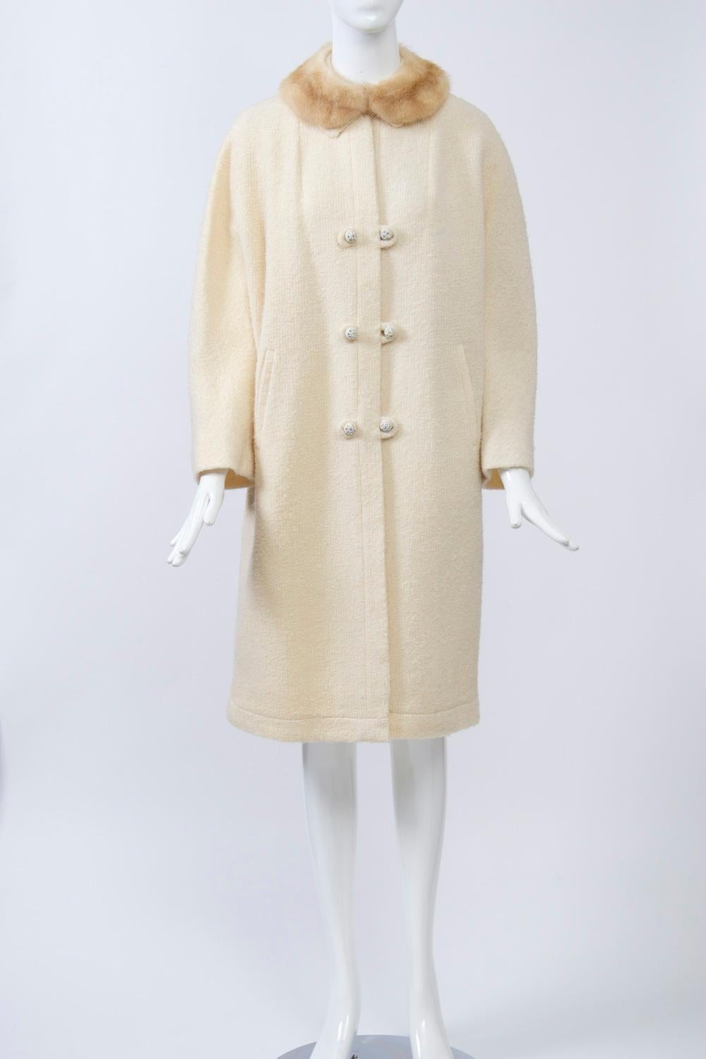 A lightweight coat from the 1960s composed of a nubby off-white wool and featuring a straight cut with dolman sleeves, a small mink collar, and a single-breasted closure system with mirror-image loops and rhinestone-studded buttons. Stitched border