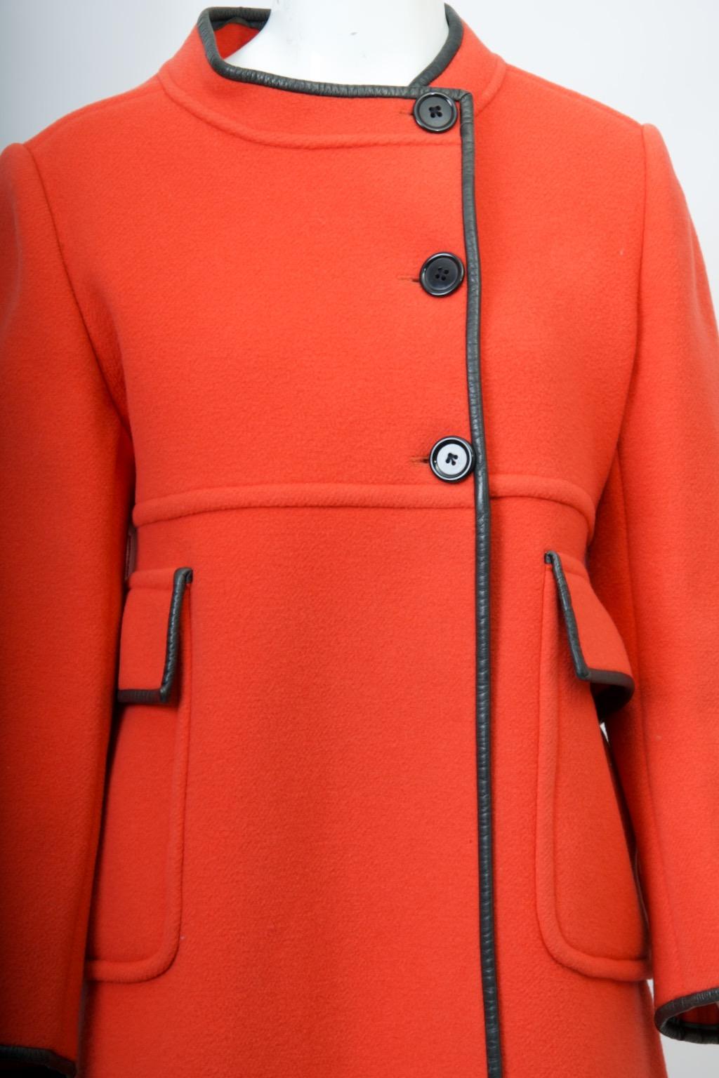 Abrigo de lana naranja de estilo icónico de los años 60, con silueta en A y costura imperio ribeteada El ribete de cuero negro en todos los bordes, incluidas las solapas de los bolsillos de imitación bajo el imperio, realza el naranja y crea