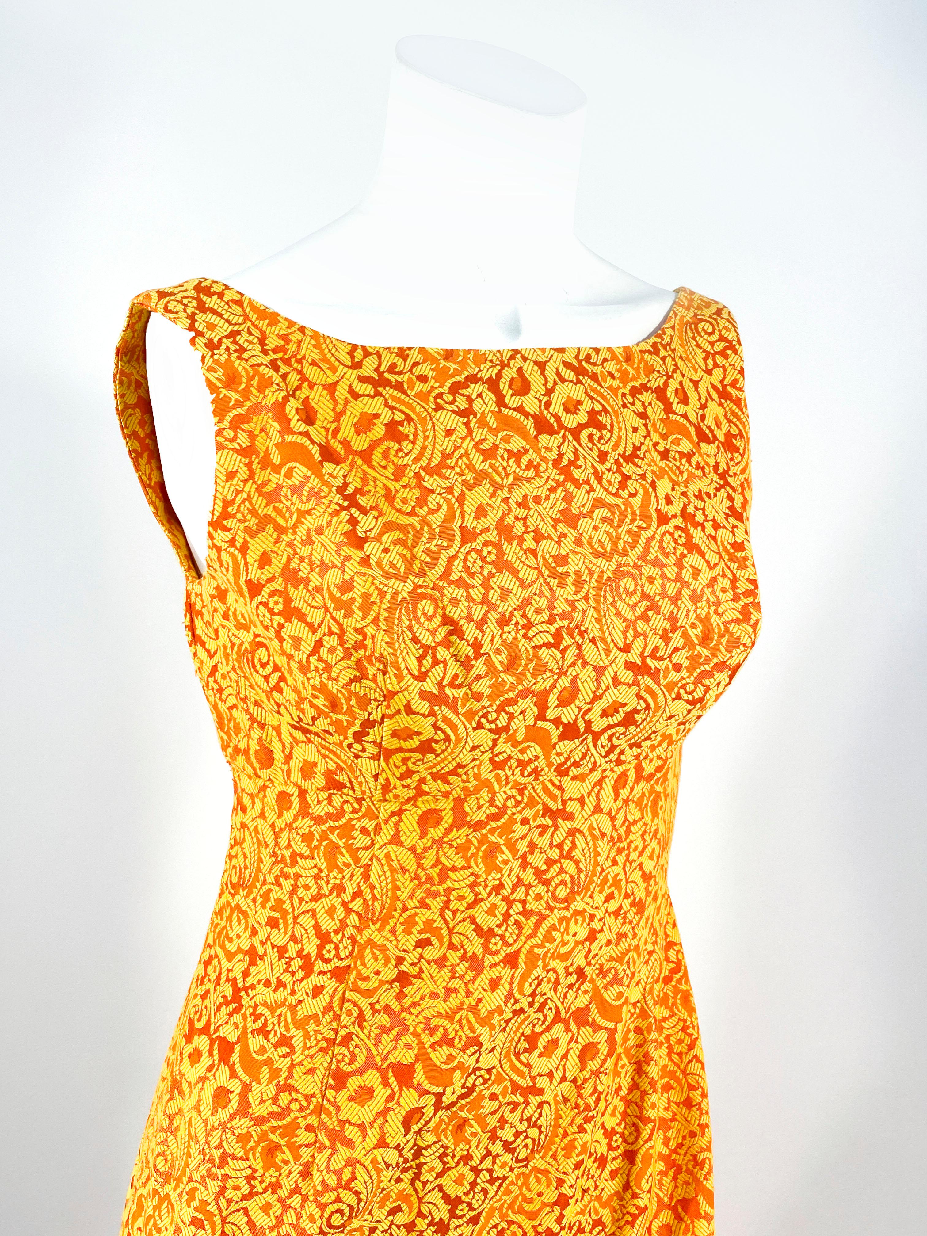 Vestido de brocado floral naranja y dorado hecho a medida, de los años 60, con corpiño imperio ajustado, falda recta y abertura trasera para mayor movilidad. El corpiño no tiene mangas y la espalda tiene un escote cuadrado sobre un cierre de