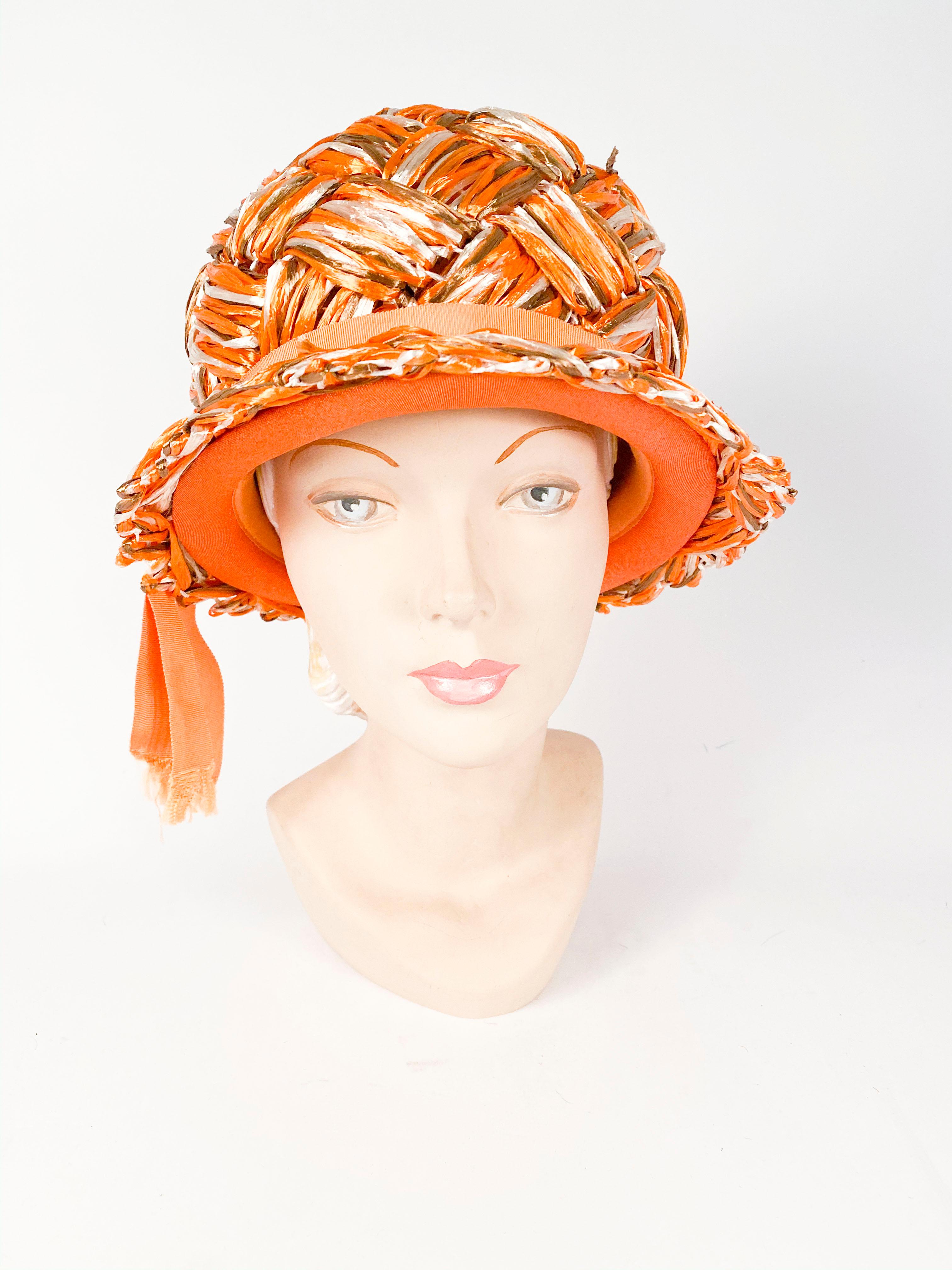 Cappello in rafia intrecciata arancione degli anni '60 con accenti marroni e beige nella trama. Questo cappello è rifinito con un nastro in grani di crescita e una coda corta con frange. 