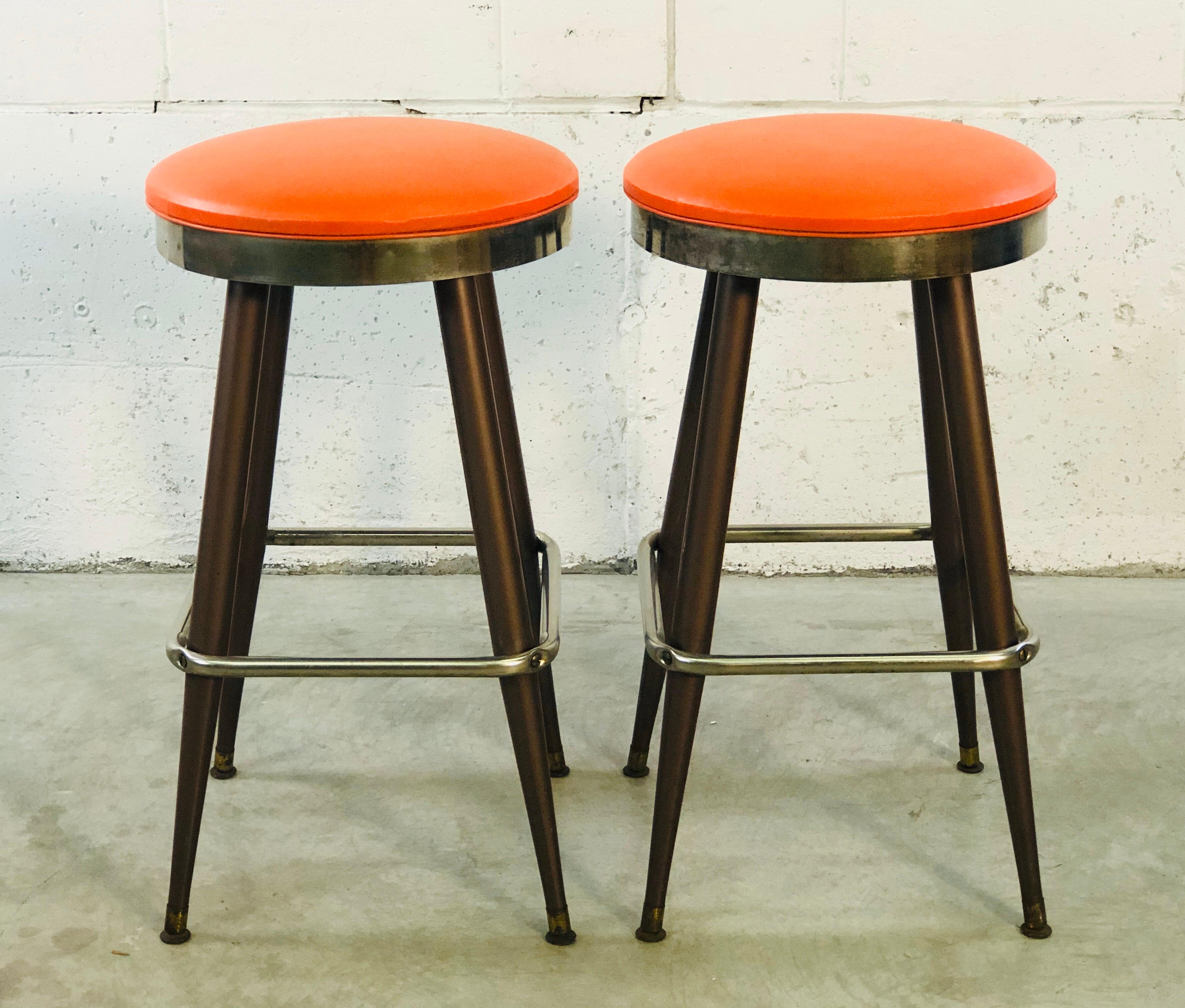 Vintage 1960s pair of orange vinyl bar stools with metal bases. The vinyl tops are round and the bases are square. Marked and dated underneath. Foot rests are 11.25” H.