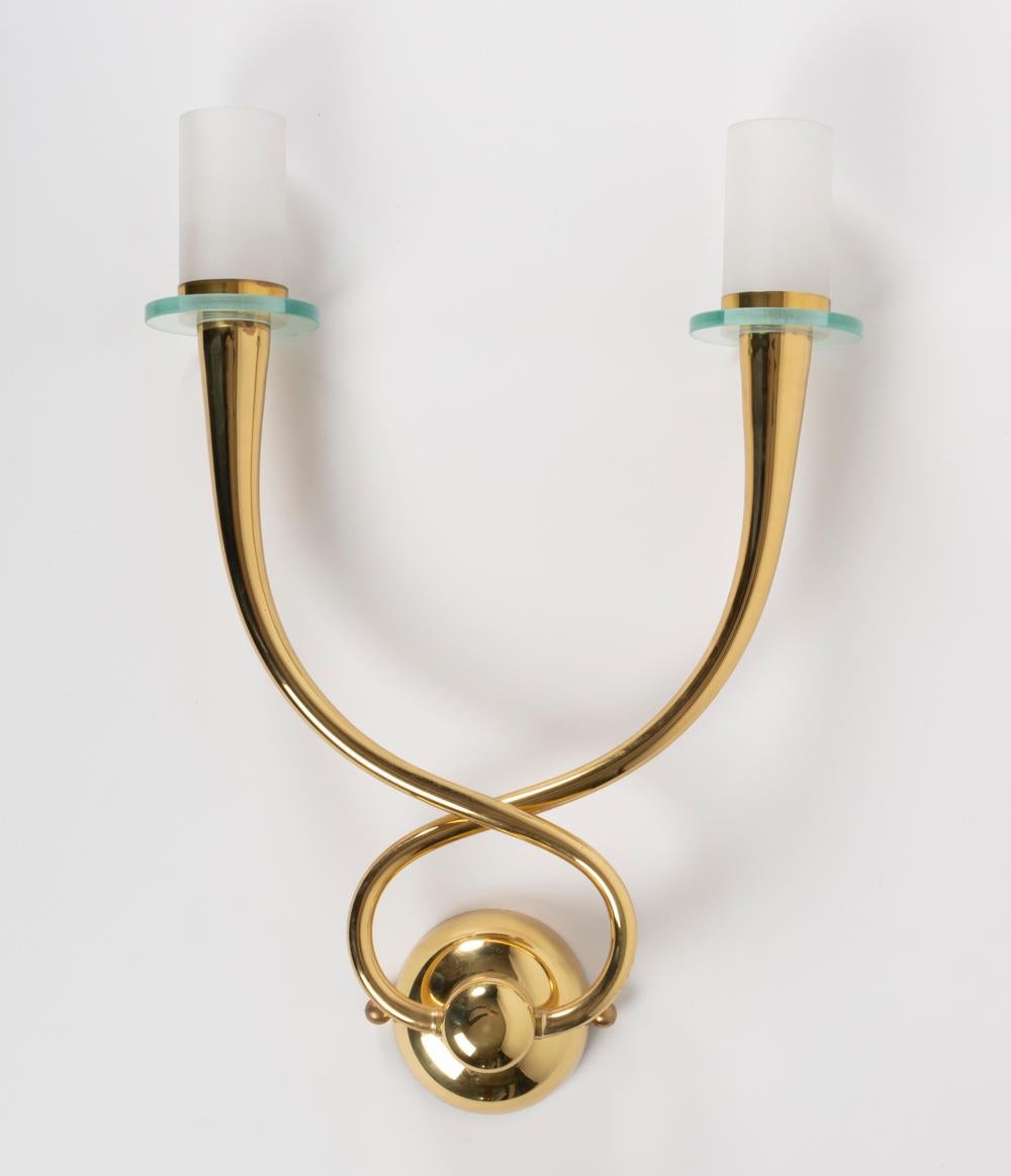 All made of brass the sconces feature two arms which intersect in the round back plate and form a buckle. The arms are ended on their upper part by glass cups and white satin glass cylinder to hide the bulb sockets.
Two bulbs per sconces.