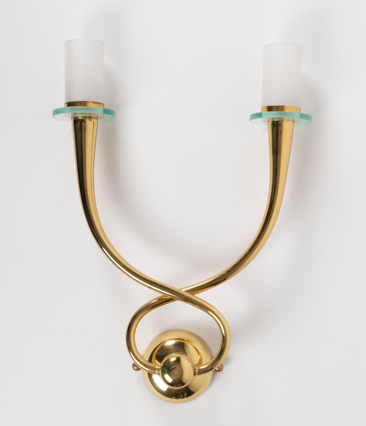 Made entirely of gilded brass, the sconces feature two stylized arms in the form of stretched cones that intersect at the base. At the intersection of the cones, a round back wall plate supports the sconce. 
On the upper part of the cones, glass