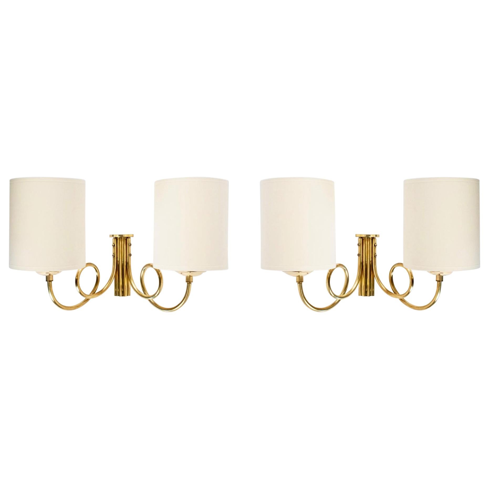 All made of gilded brass.
The two lighted arms are bend to form a pair of buckles. 
The back plate is a fluted half cylinder.
Cylindrical off-white cotton shades.

Two bulbs per sconces.
