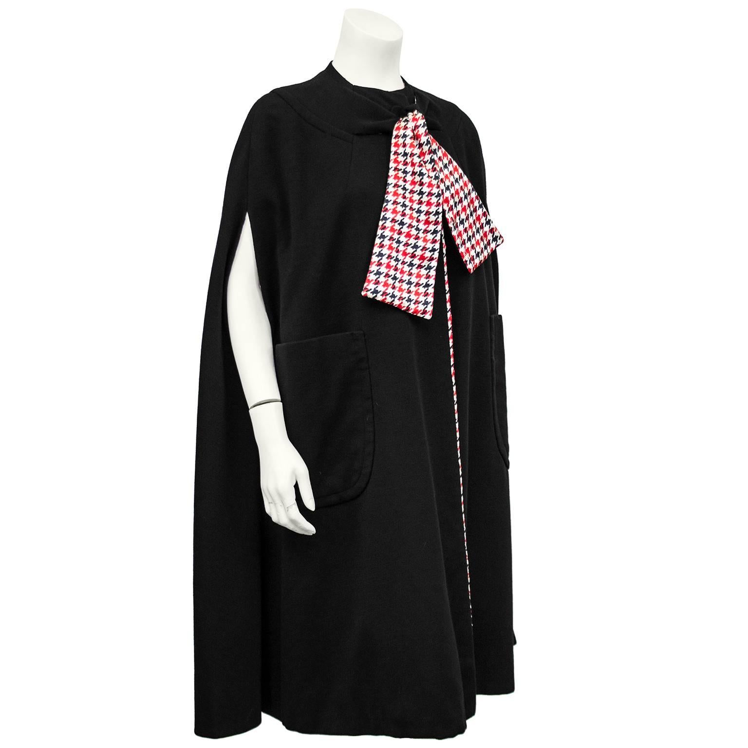 1960s Pauline Trigere black wool jersey cape. Red, black and cream houndstooth silk lining for a pop of colour. Large pussy bow tie at neck with black on one side and houndstooth lining on the other - when tied the houndstooth faces out. Adorable
