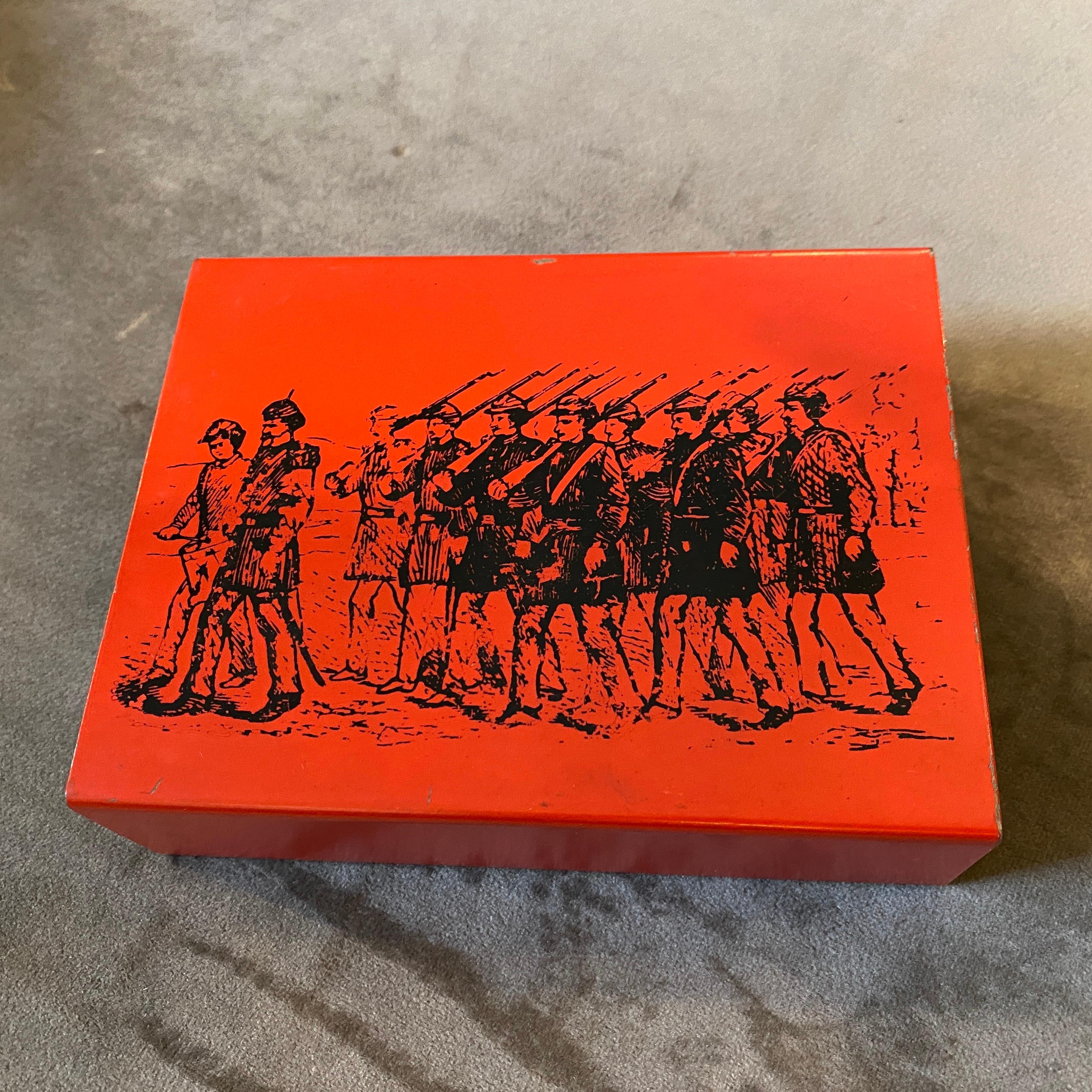 A mahogany and red metal box with black decor depicting soldiers. It's designed by Piero Fornasetti and manufactured by Atelier Fornasetti. The Cigarette Box is a highly collectible and distinctive piece that showcases the iconic style of Piero