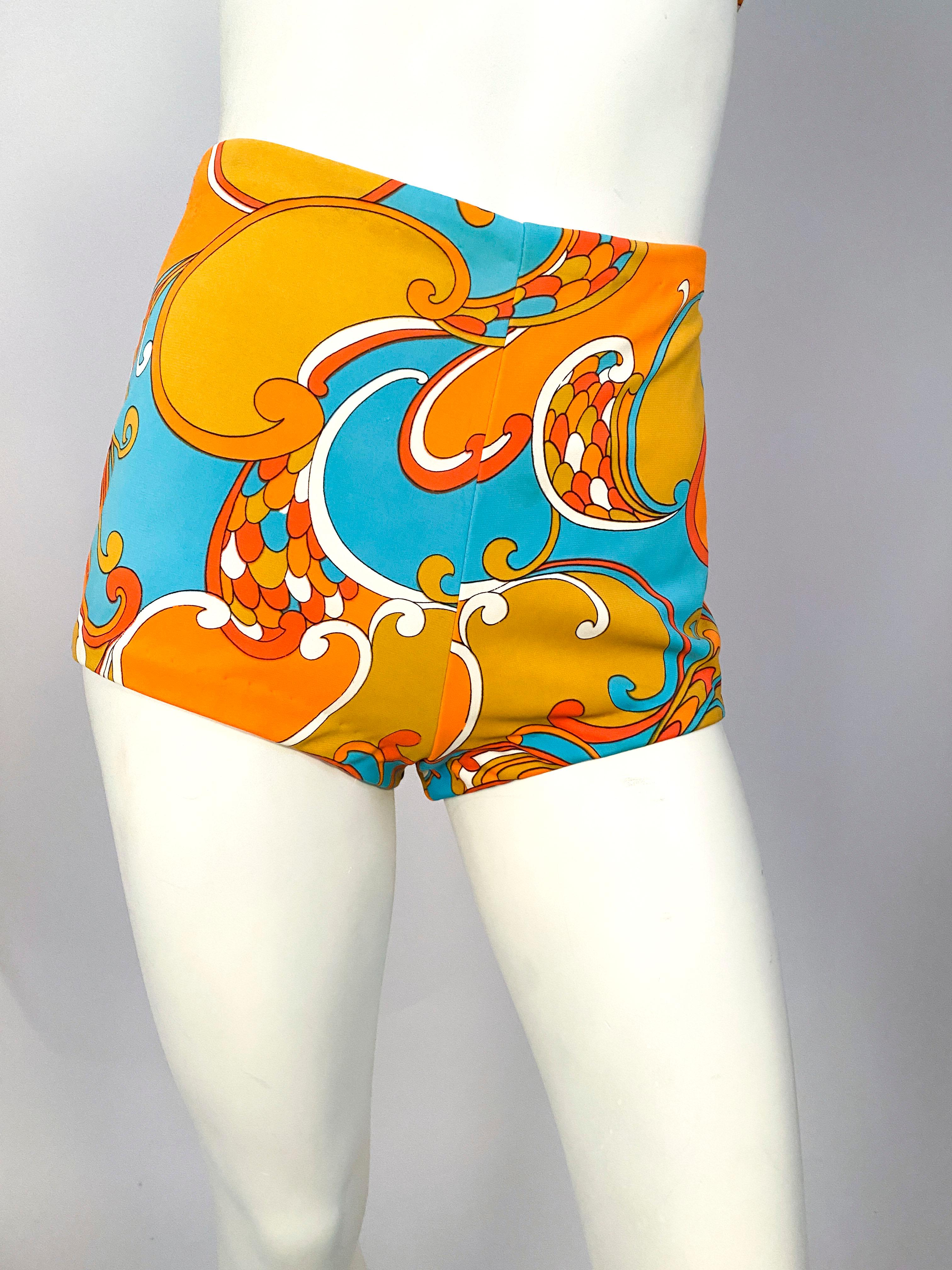 1960s two-piece bathing suit featuring a psychedelic (Pucci -esque) print in neon orange, aqua, tangerine, white, ang gold. The top has a halter structured strap that buttons to the back of the bust strap. The bottoms are high waisted and