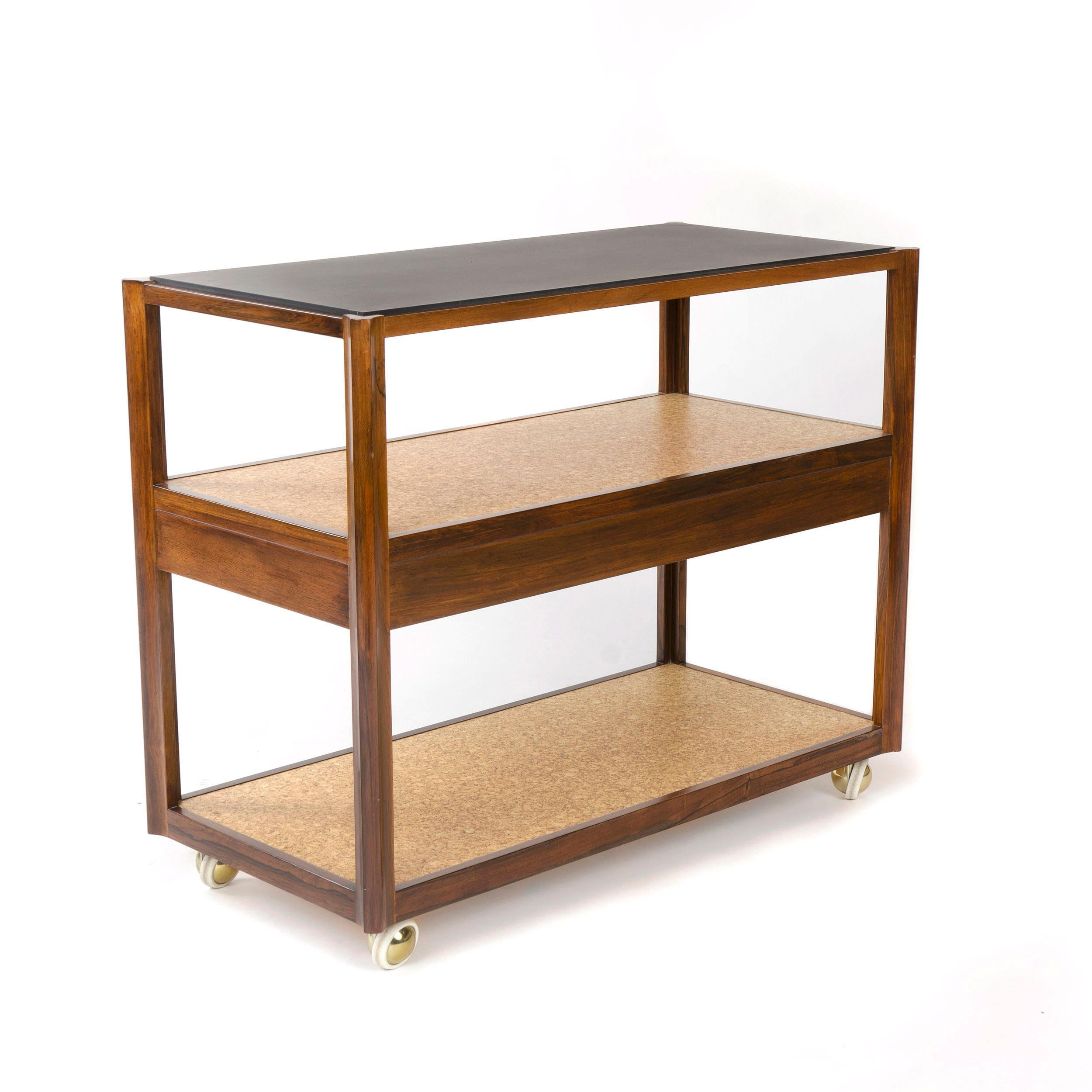 Rosewood framed three tier service or utility cart. The top tier has an inset slate top above two cork shelves with the middle shelf having a blind pull suspended drawer for miscellaneous storage. The frame is mobile via four Shepard ball castors in