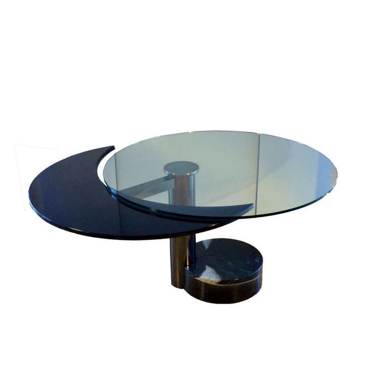 Sculptural dining table revolves in two positions from oval to round. When the black lacquered part is moved down and turned under glass tabletop the table becomes round. When extended the black section lifts up to the glass level. 
The foot is in