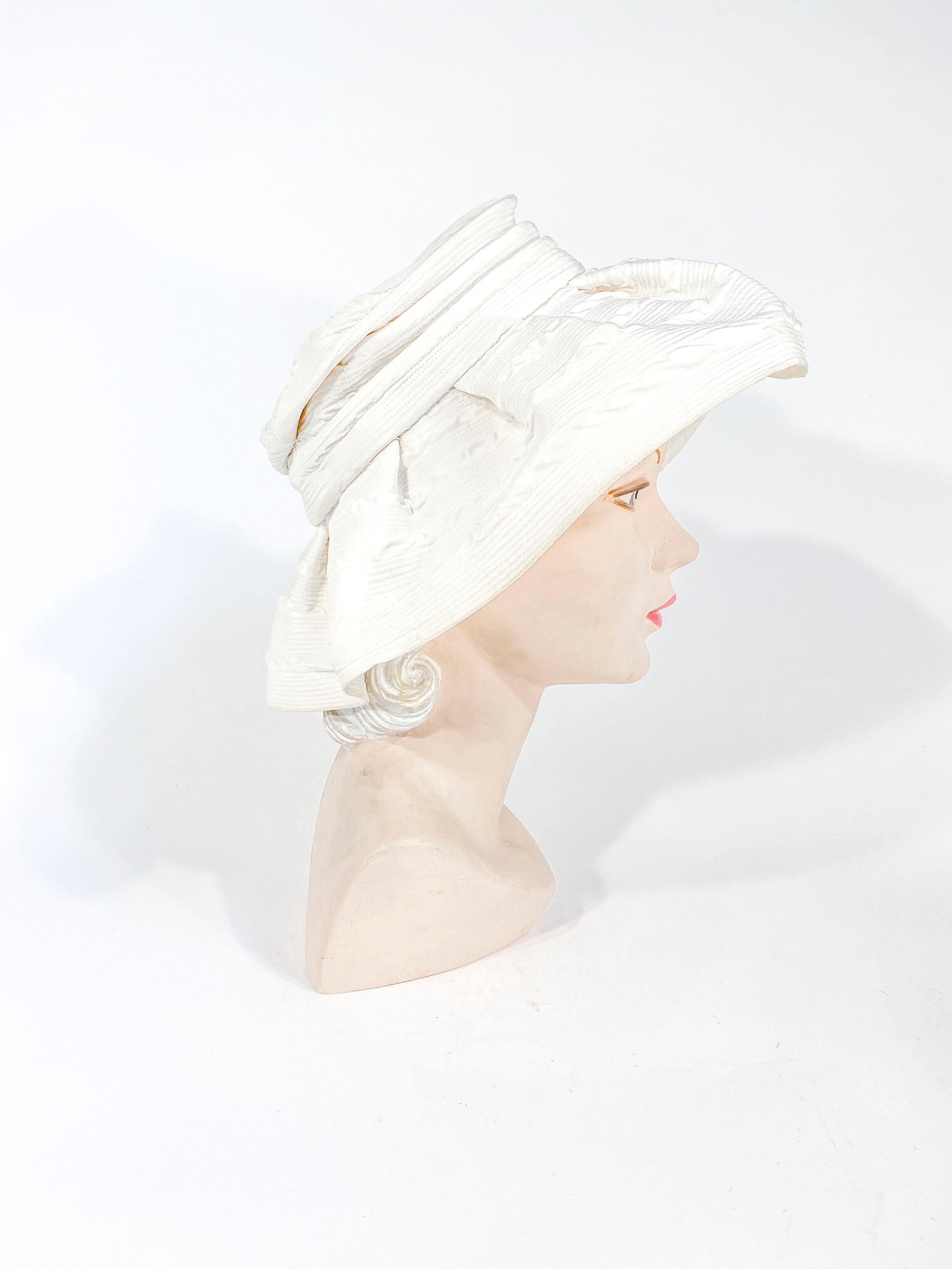 Chapeau de mode blanc cassé des années 1960 avec un large bord non structuré froncé sur la couronne. Le textile de till présente de fines surpiqûres circulaires qui enveloppent le chapeau. 