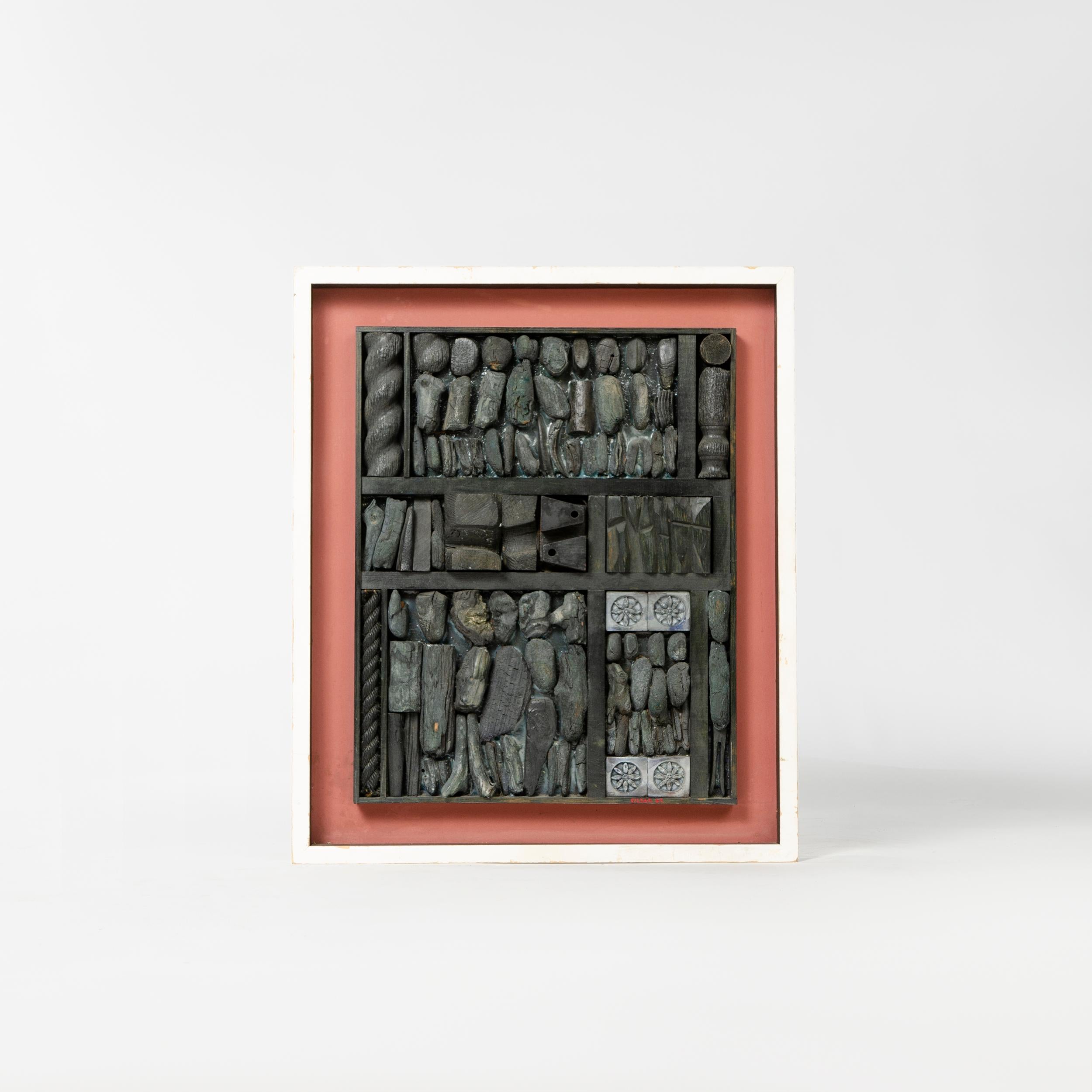 A blackened sculptural relief collage by William Sildar with dark pink matte backdrop and white painted wood frame.