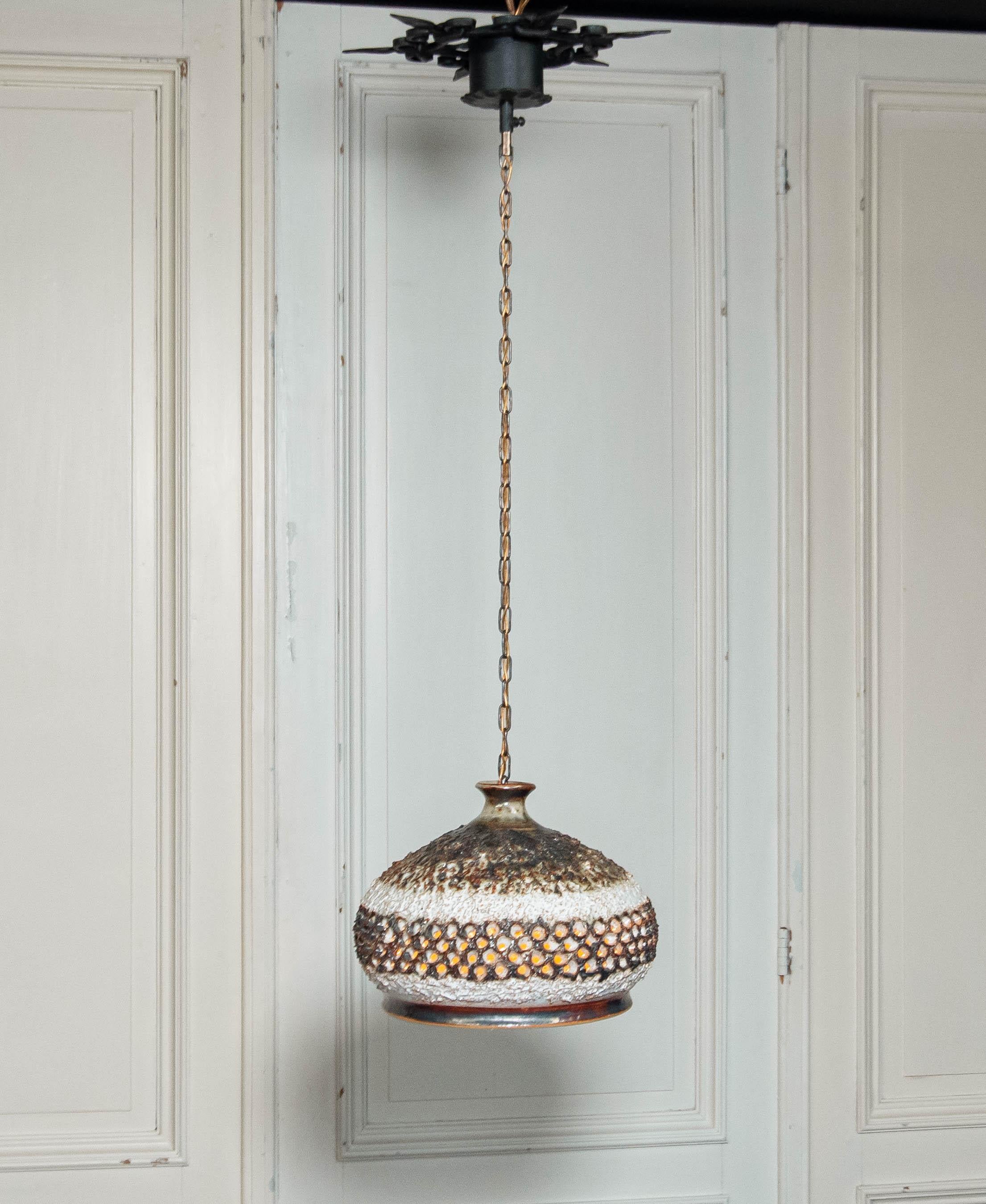 Questa sorprendente lampada a sospensione brutalista degli anni '60, prodotta dalla fabbrica Sejer, mette in mostra:

Un paralume in ceramica lavorato a mano con una superficie grezza e strutturata
Il design traforato crea un effetto di luce