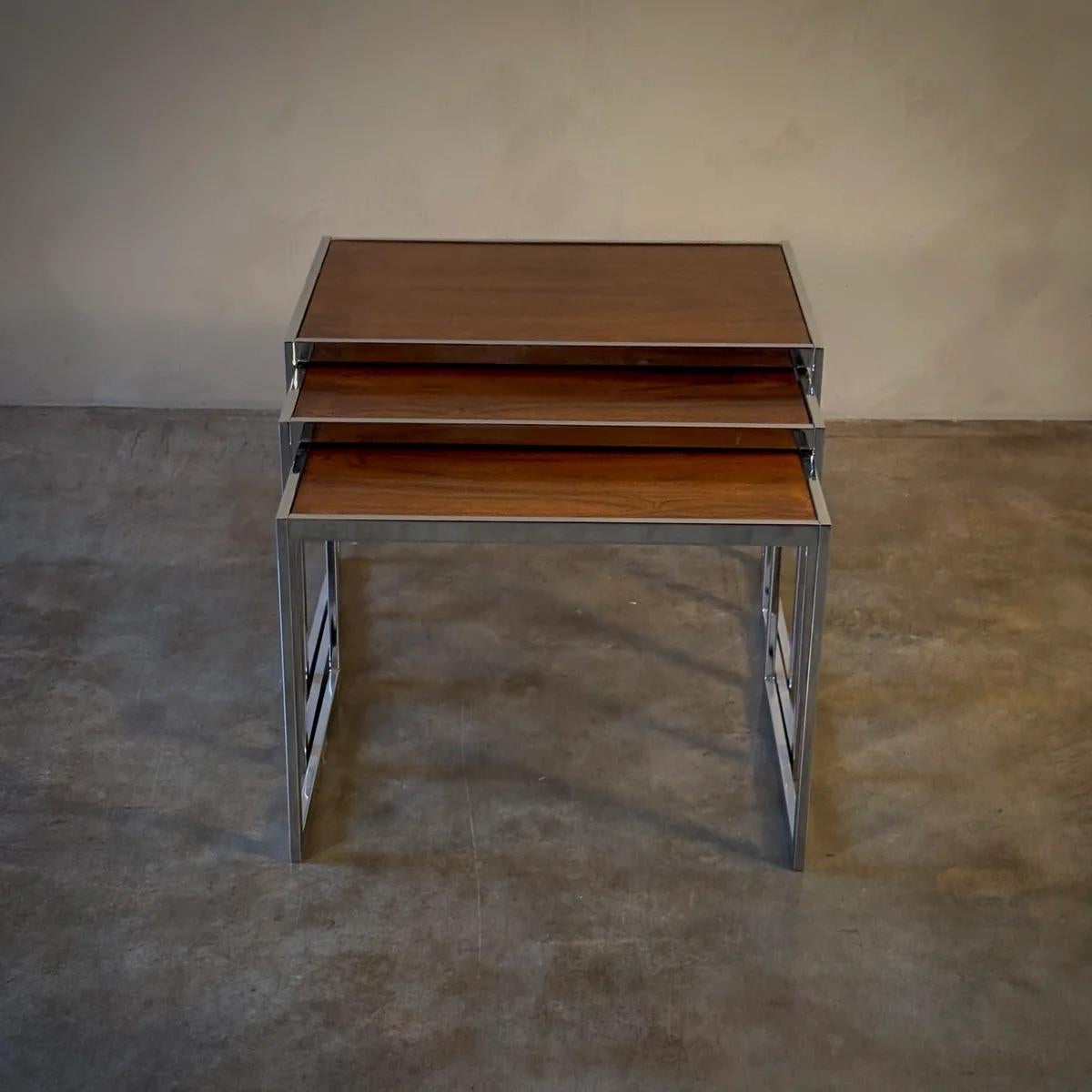 A sleek assembly of functionality and style, this set of three English mid-century nesting tables showcases the perfect blend of wood and chrome. Each table, varying slightly in size, offers practicality without sacrificing visual appeal. The
