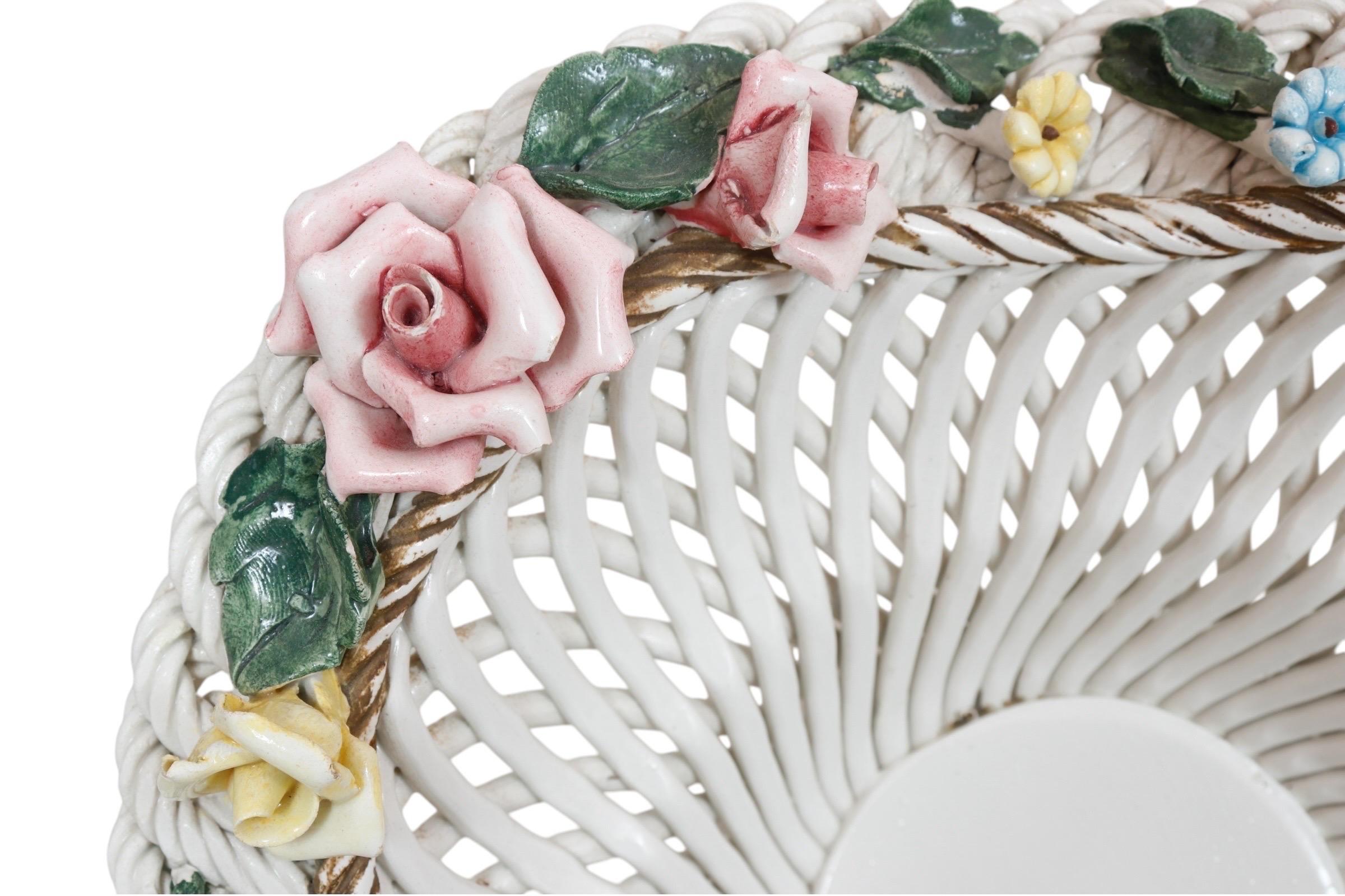 Una fruttiera in ceramica firmata Bassano del 1960 circa. Un cesto a reticolo traforato con lati ondulati è decorato con un bordo intrecciato a corda e fiori in giallo, blu e rosa. Il piede è decorato con volute a c e un motivo di foglie d'acanto.