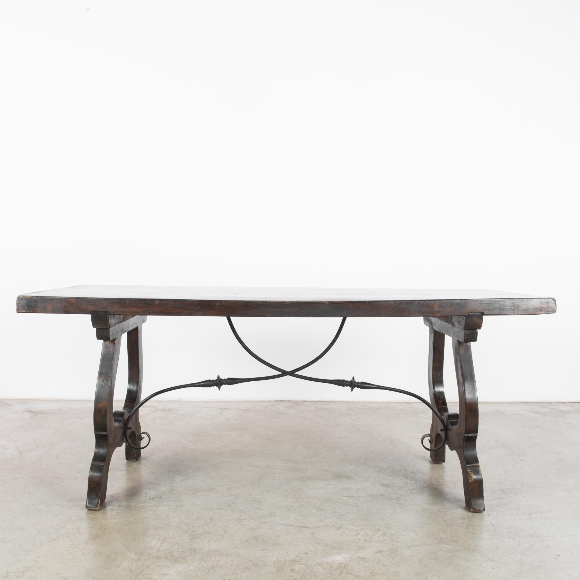 A wooden dining table from Belgium, circa 1960. Lyre-shaped legs support the broad slab of the tabletop. The dark wood has a rich, burnished tone. Beneath the tabletop, curving cast iron tendrils swoop and intersect in romantic embellishment,