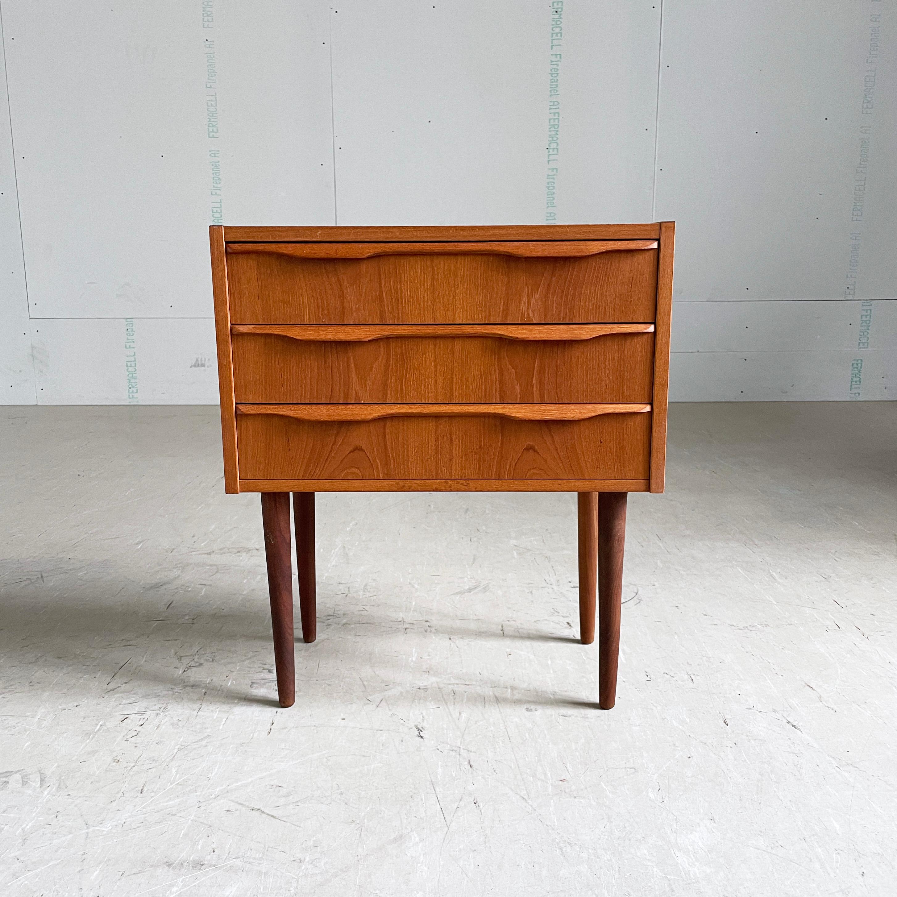 Mid Century teak bedside cabinet / chest of drawers. Teak veneer with solid teak legs. Three identical sized drawers with beautiful detailing to handles. 
Origin unconfirmed, possibly Danish. Produced ca. 1960. 
