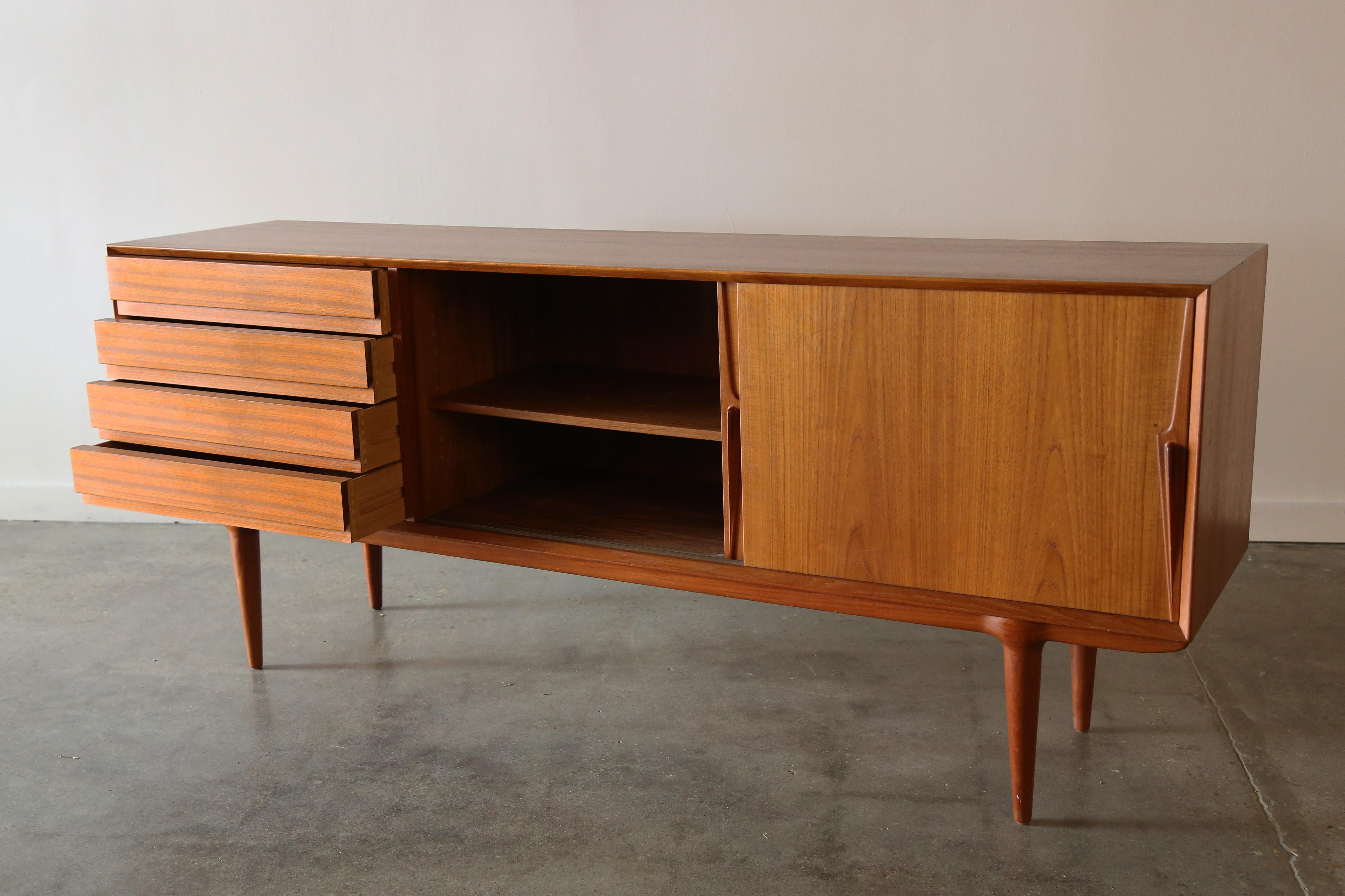 A sculptural teak credenza designed by Gunni Omann. An uncommon form, Model 18 features two sliding doors with solid teak sculpted handles, concealing a shelved interior. A row of drawers on the left side with recessed pulls, all floating on top of