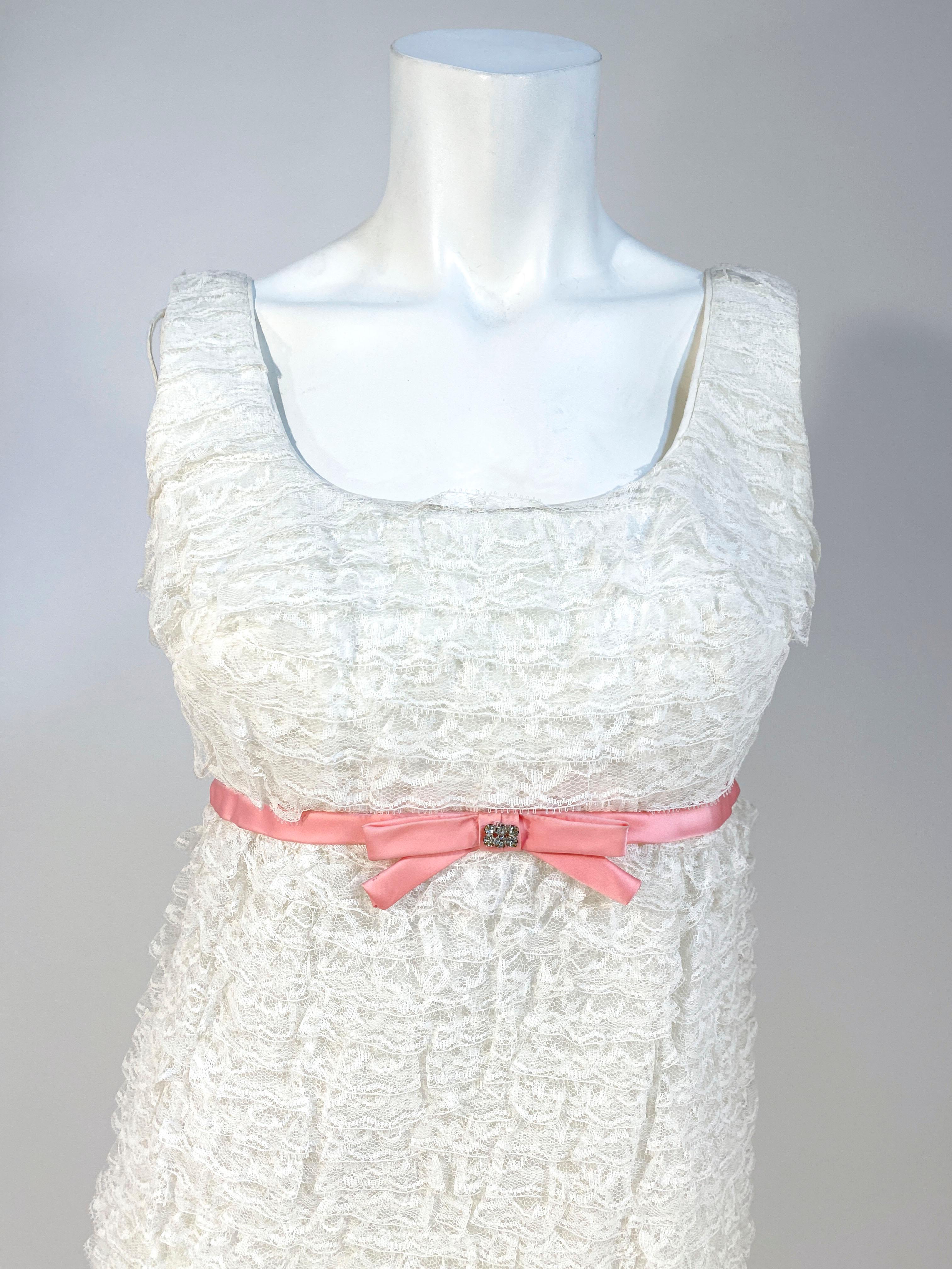 1960s party dress with multi-rows of off-white lace from the top of the shoulders to the skirt hem. The empire was it is decorated with a pink heavy stain piping finished with a bow and rhinestone accent. 