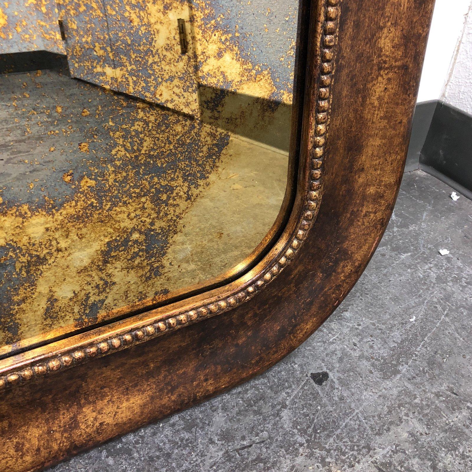 Aa wood frame framed wall mirror. The frame is wood with a nailhead trim border with curved ends in a distressed gold finish. The mirror has an antiqued finish. Wall mirror hangs horizontal.
 
