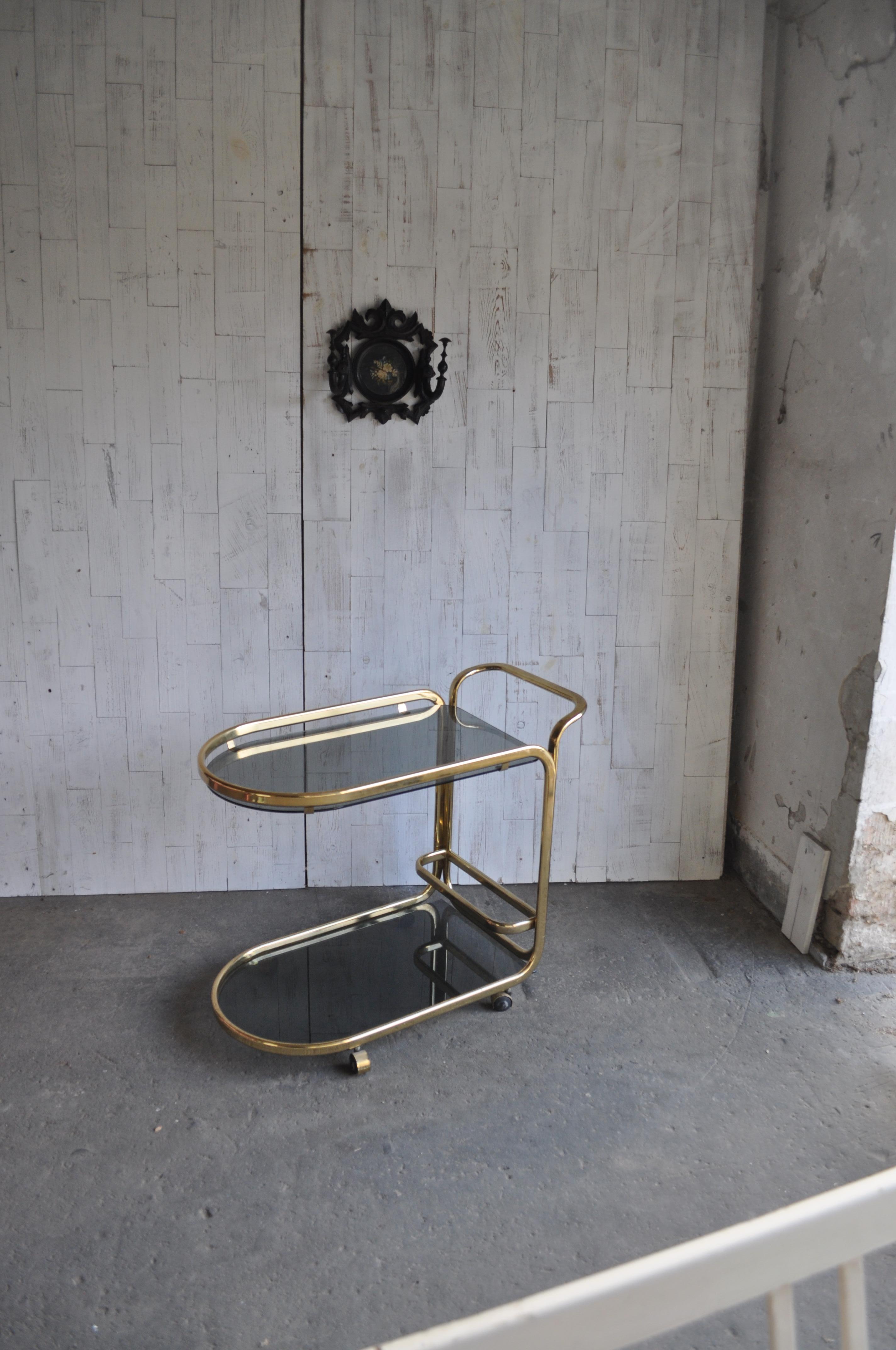 This 1960s Vintage French Regency-style tea cart a brass frame. The brass frame features two levels. The top level features a glass shelf with a bar around the top with a handle to push the cart. The cart is finished with round caster wheels for