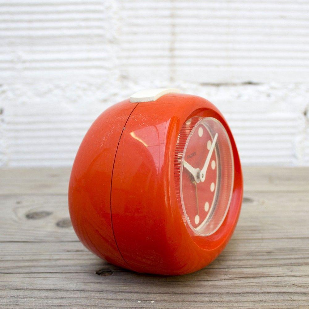 This tabletop clock is from the 1960s and manufactured by the German brand Kaiser. The plastic structure is a red orange color with white detailing on the clock face and hands. Its rounded lines and clock face design have are of a Mid-Century Modern