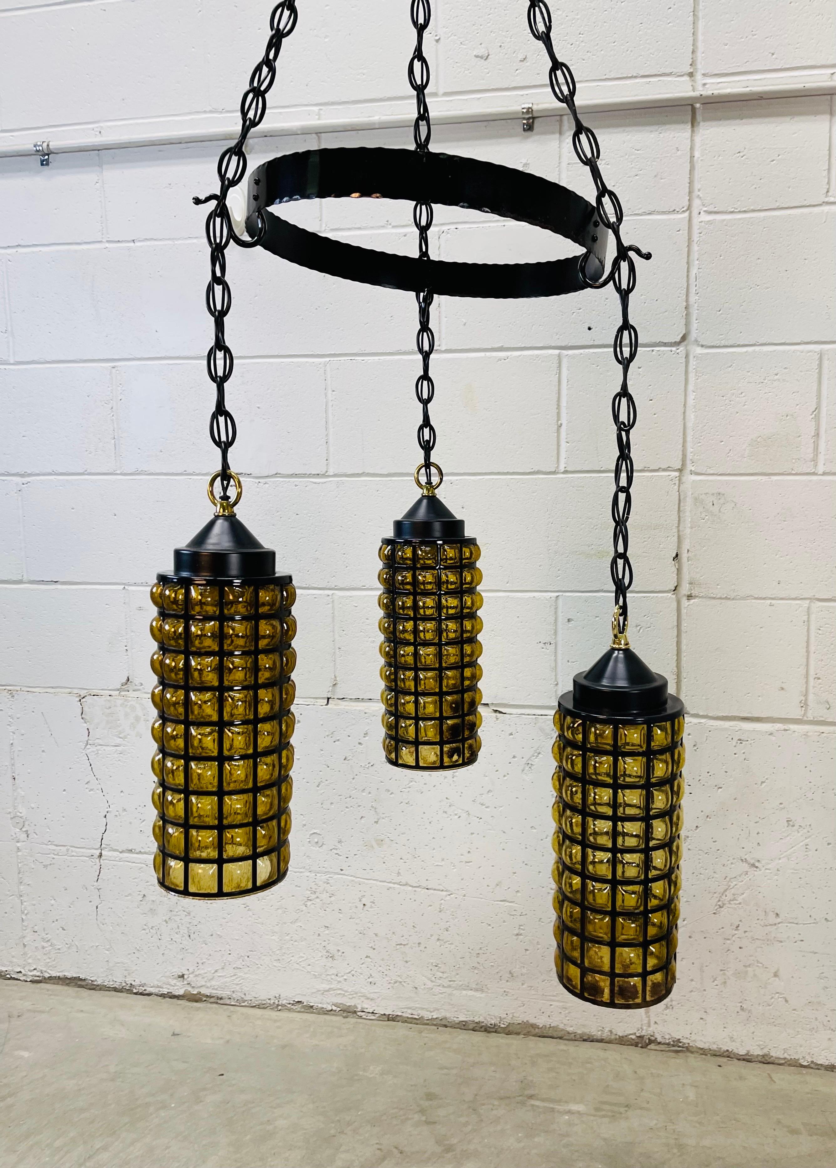 Vintage 1960s black metal and handblown amber glass pair of pendant lights (only one shown). The lights are hardwired and will need to be assembled. The three pendants hang from the round metal center. Chain drop is 22”H max and can be adjusted. The