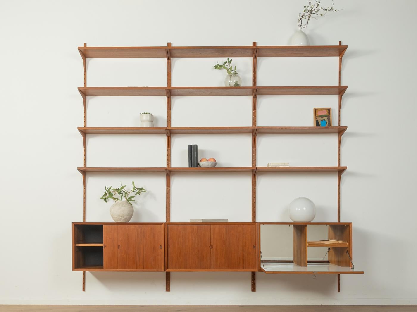 Classico sistema di scaffalatura degli anni '1960. I contenitori e i ripiani di alta qualità sono impiallacciati in teak. Il sistema è composto da dodici ripiani, un contenitore a barre con porta a battente e cassetto interno, due contenitori con