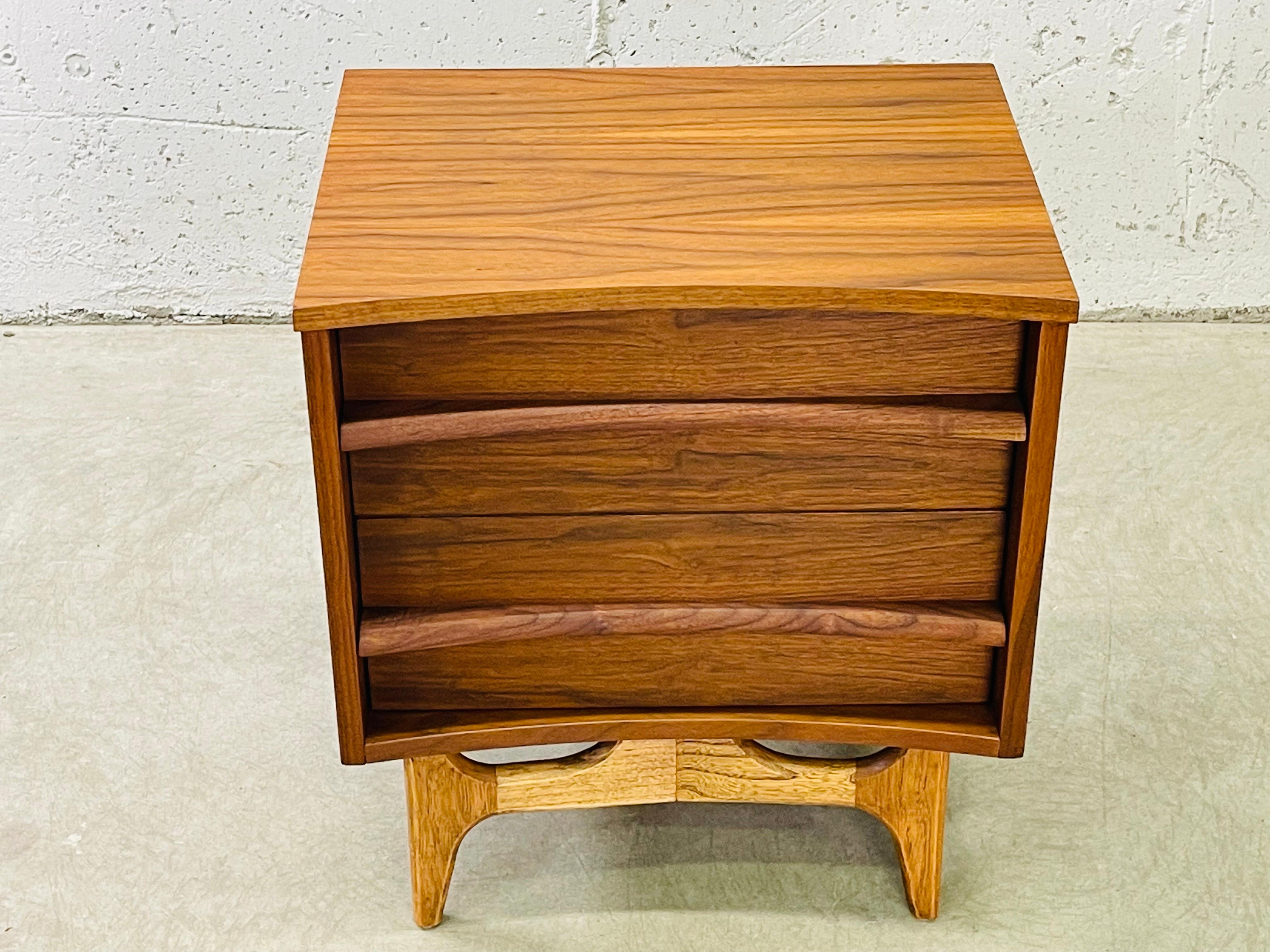 Vintage 1960s walnut wood curved front two drawer nightstand. The nightstand sits on an ashwood base. The drawers have a walnut wood pull. Newly refinished condition. No marks.