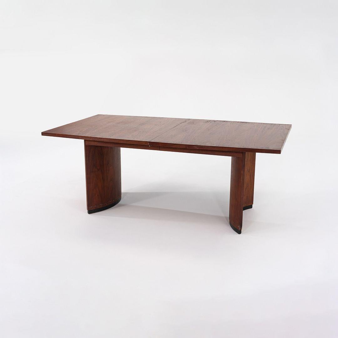 This is an Extension Dining Table in Walnut, dating to the 1960s. It is attributed to Jack Cartwright for the manufacturer Founders. The design features a curved slab base. The piece is formed of solid and veneered walnut and is accompanied by two