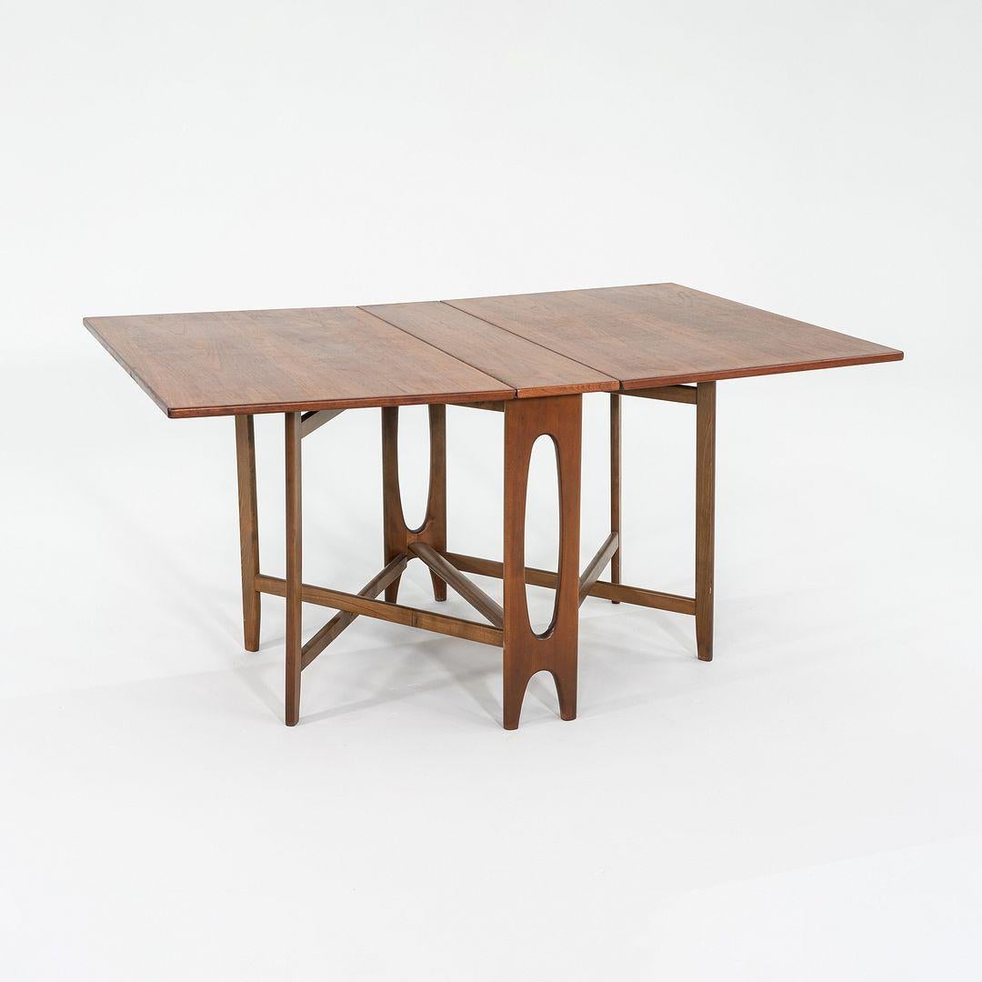 Se trata de una mesa de comedor con patas de nogal, diseñada por Bendt Winge en los años 50 y fabricada por Kleppe Mobelfabrikk en Noruega. Está hecha de madera de teca, e incorpora una hoja a cada lado que amplía la anchura de la mesa cuando está