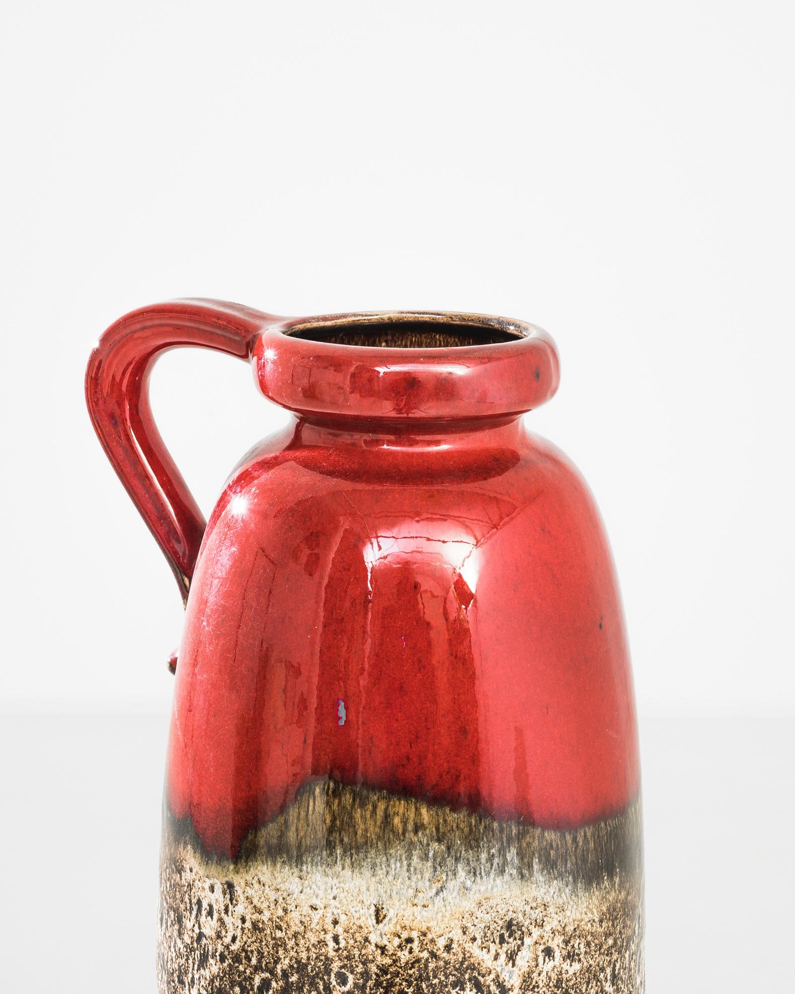 This vibrant 1960s West German ceramic vase is a splendid example of mid-century modern design. Its bold red glaze, which smoothly transitions into a speckled natural finish, captures the eye and evokes a sense of dynamic contrast. The sleek,