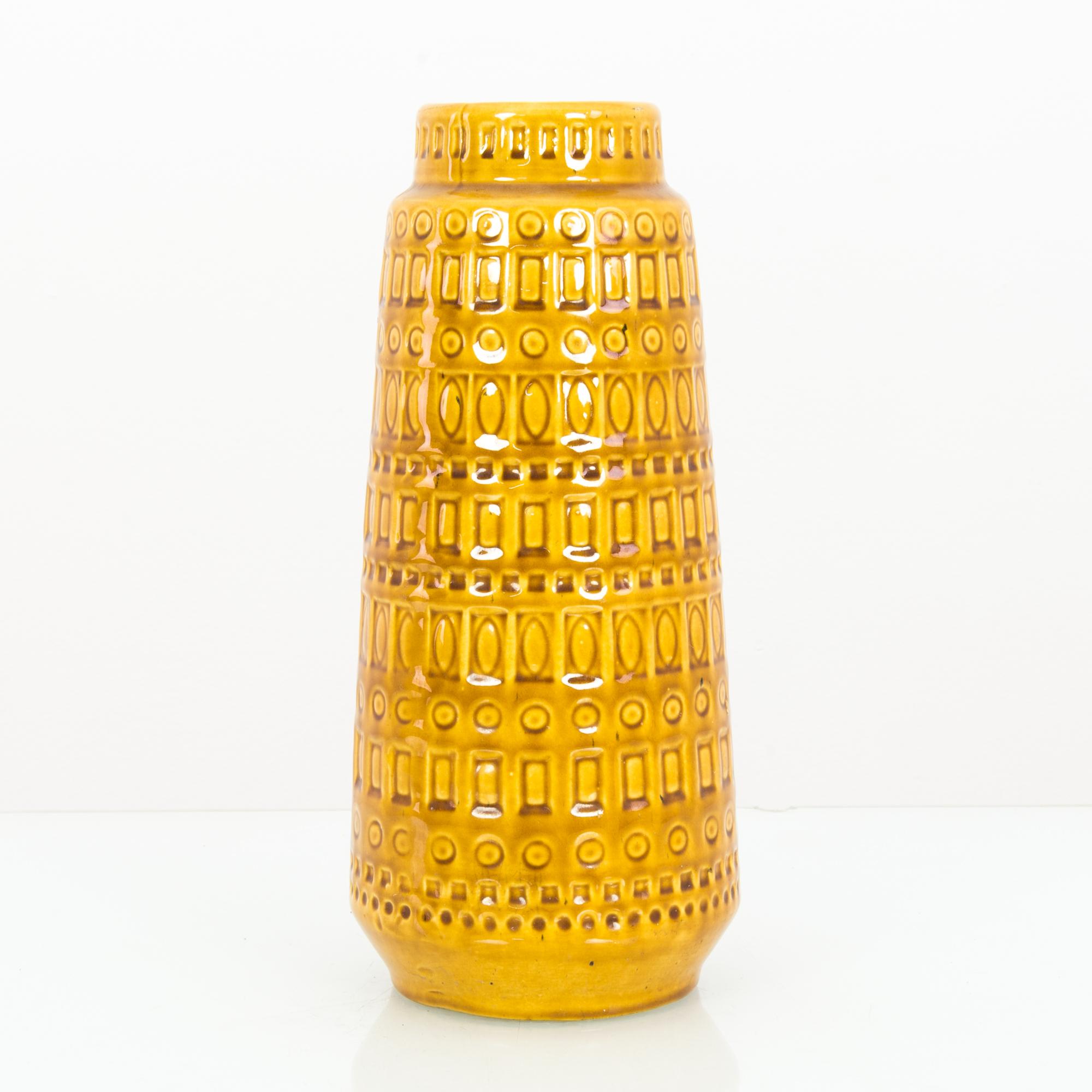 A ceramic vase from West Germany, circa 1960. A tall, tapering form, decorated with geometric impressions. The glaze is a goldenrod color, bright yet earthy, with a cheerful gleam. The shape is reminiscent of a circular tower of babel, girded with
