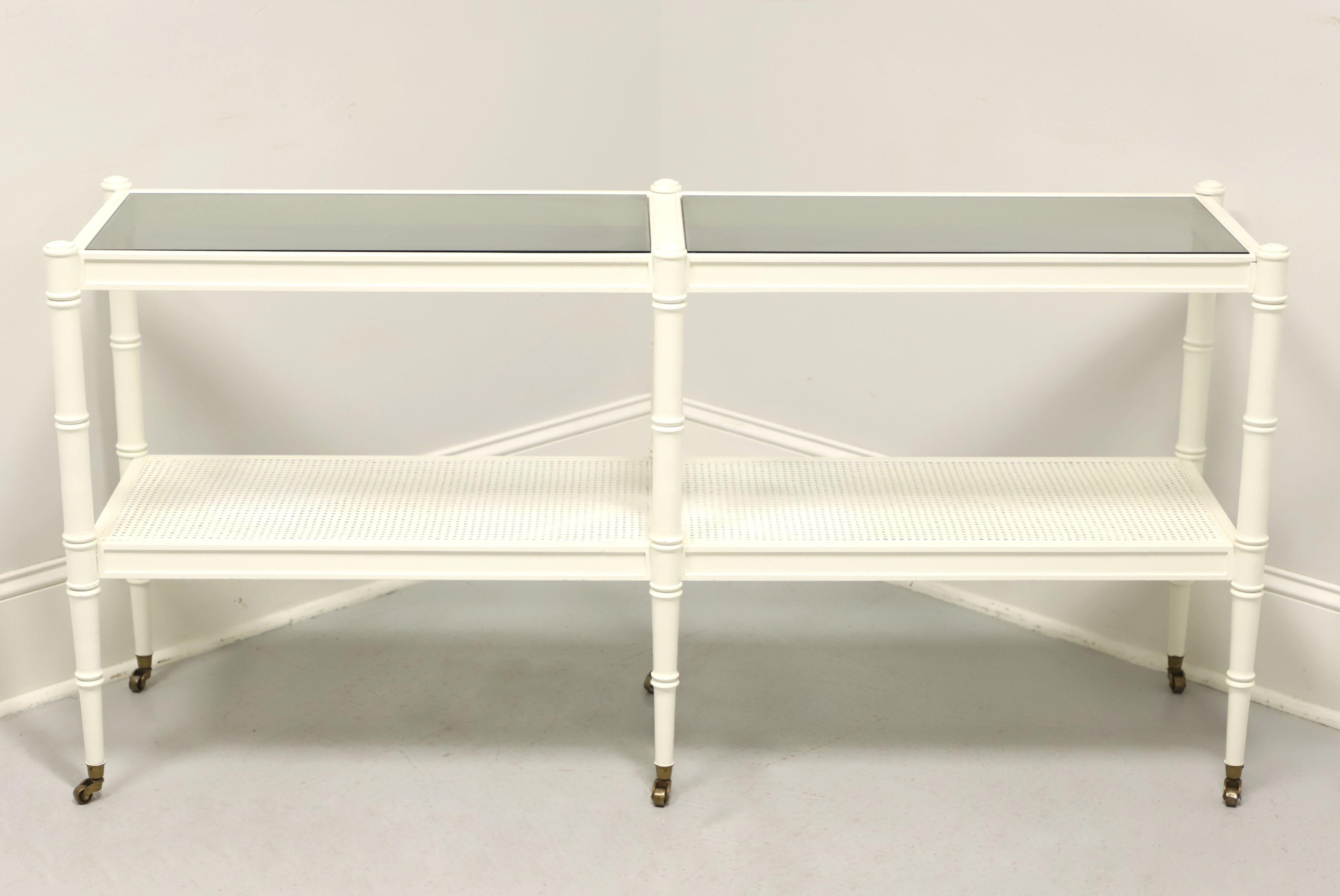 A Hollywood Regency style console table, unbranded, similar quality to Tomlinson Furniture. Faux bamboo painted white, an upper tier with dual smoky black glass top, lower tier being caned, round tapered legs and six brass caster. Made in the USA,