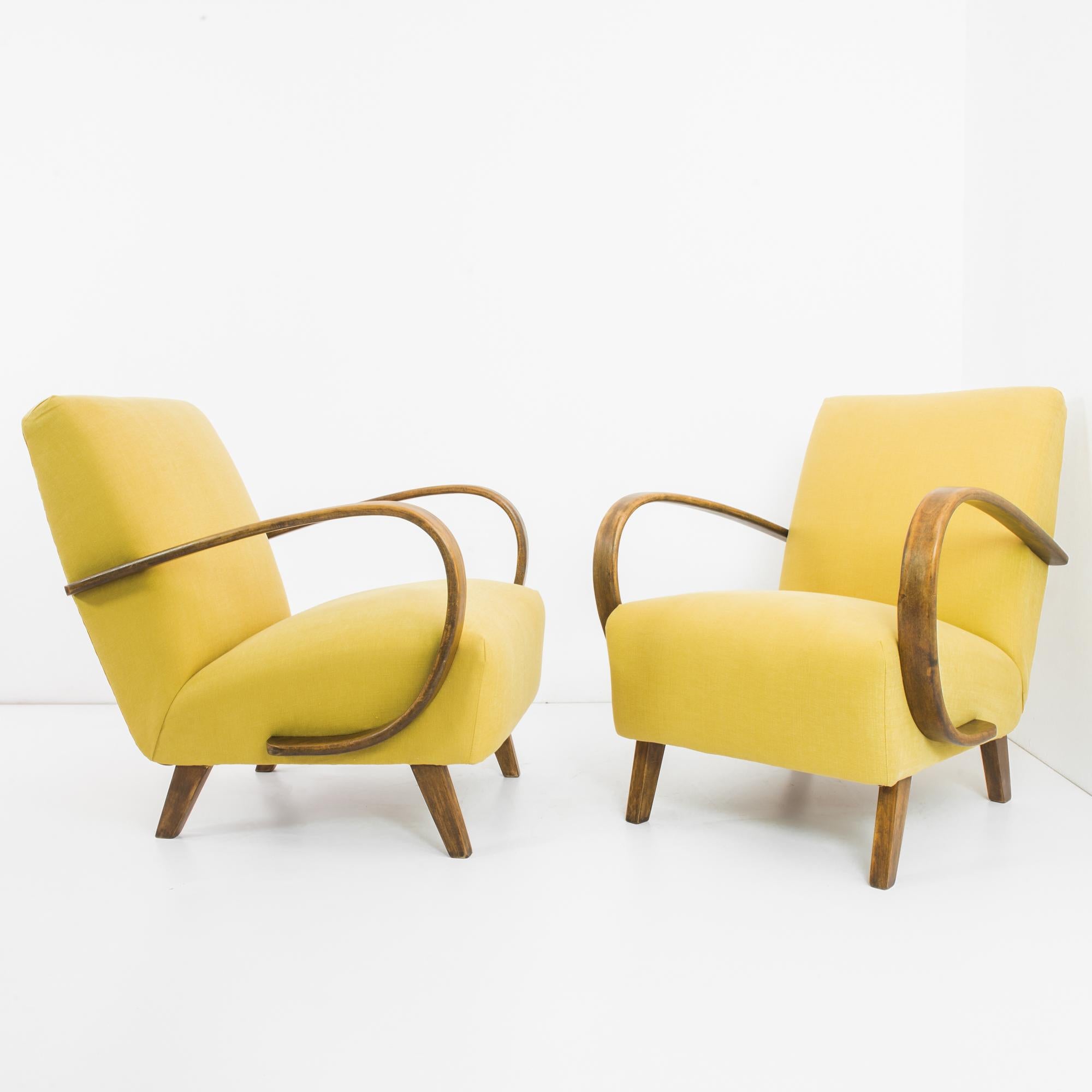 A pair of 1960s armchairs by Czech furniture designer J. Halabala. A low-set, reclined silhouette, upholstered in a lemon yellow fabric. The deep cushioned seat is braced by the distinctive sweep of the polished bentwood armrests. The form is