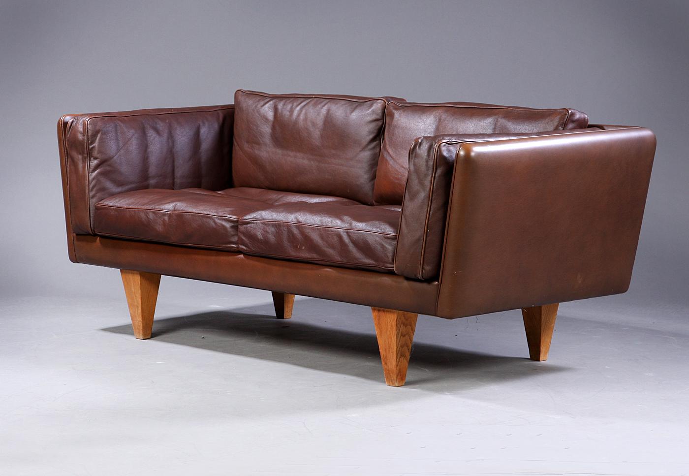 Illum Wikkelsø 1919-1999. Two-seat sofa, freestanding, upholstered in brown patinated sturdy leather, loose cushions with down filling, cone-shaped oak legs. Produced by Holger Kristiansen, V11 series, 1960s-1970s. Measures: H. 67/41 cm. L. 167 cm.