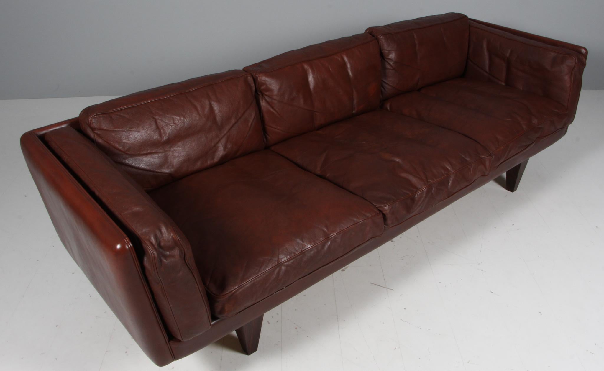 Illum Wikkelsø 1919-1999. Three-seat sofa, freestanding, upholstered in brown patinated leather, loose cushions with down filling, cone-shaped rosewood legs, 1960s 1970s. Produced by Holger Kristiansen, V11 series. 

